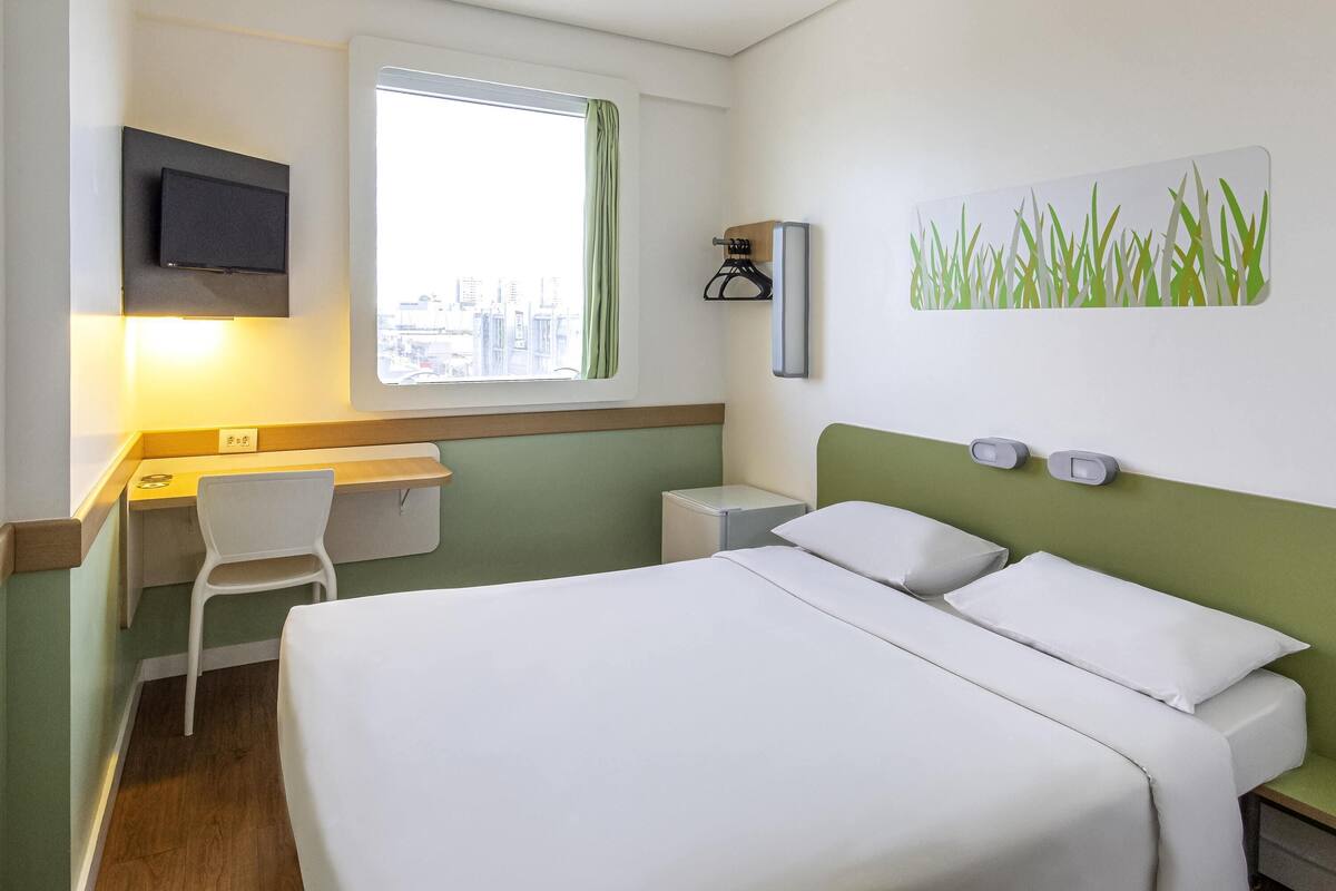 A hotel room with a bed, a desk, a chair, a TV, and a window with a curtain.