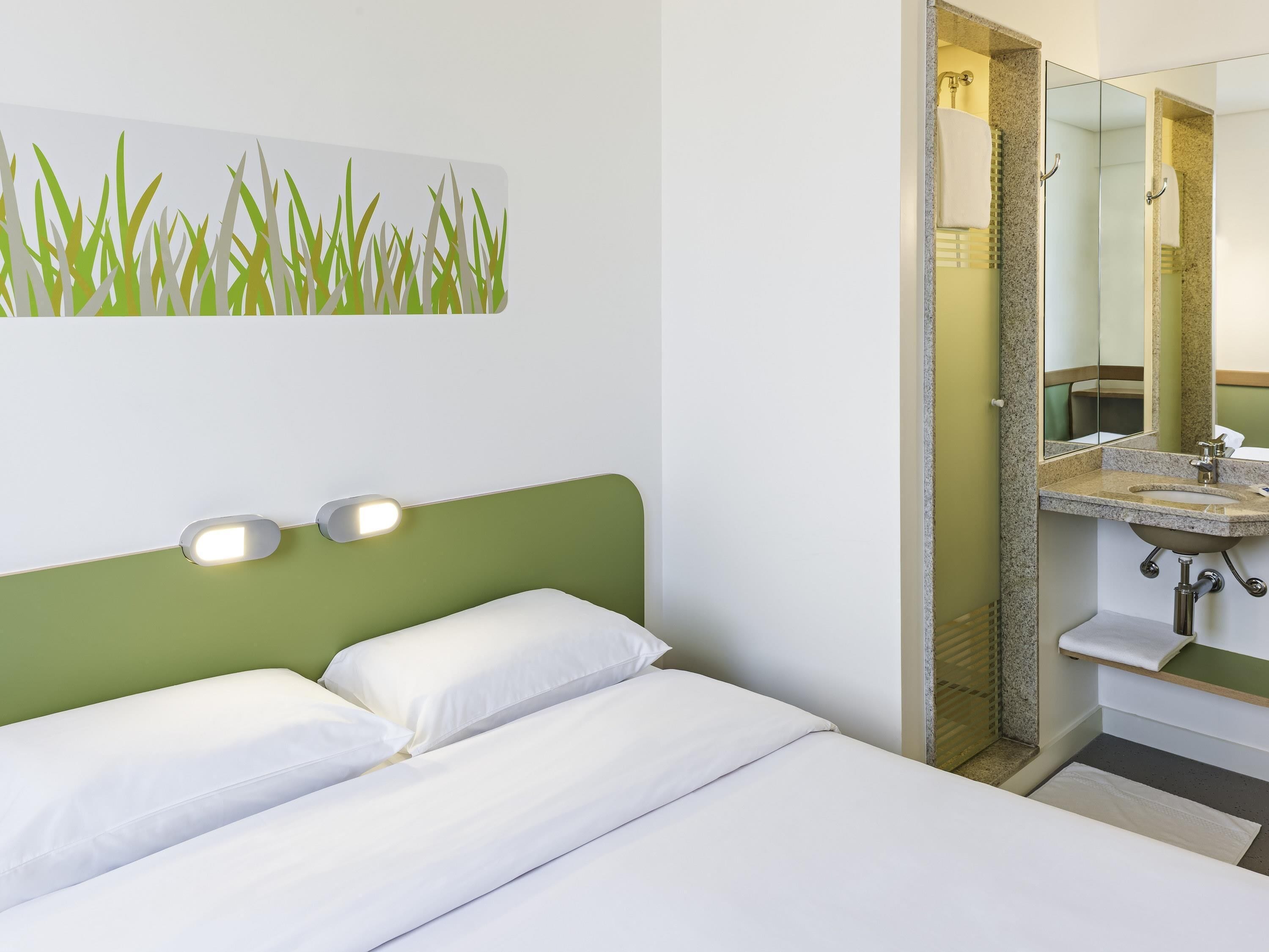 A hotel room with a bed, a green headboard, a wall-mounted light, and a bathroom with a sink and mirror.