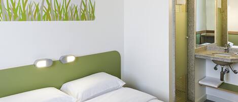 A hotel room with a bed, a green headboard, a wall-mounted light, and a bathroom with a sink and mirror.