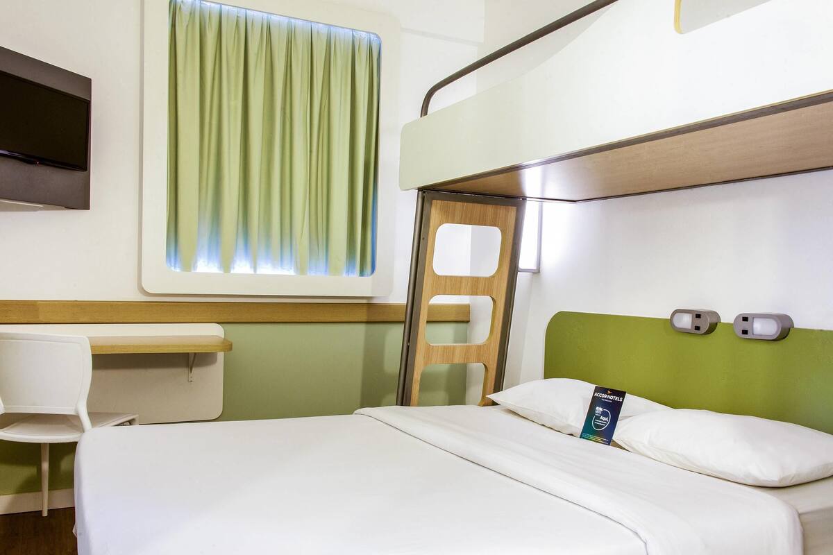 A hotel room with a bed, a desk, a chair, a TV, and a window with curtains.