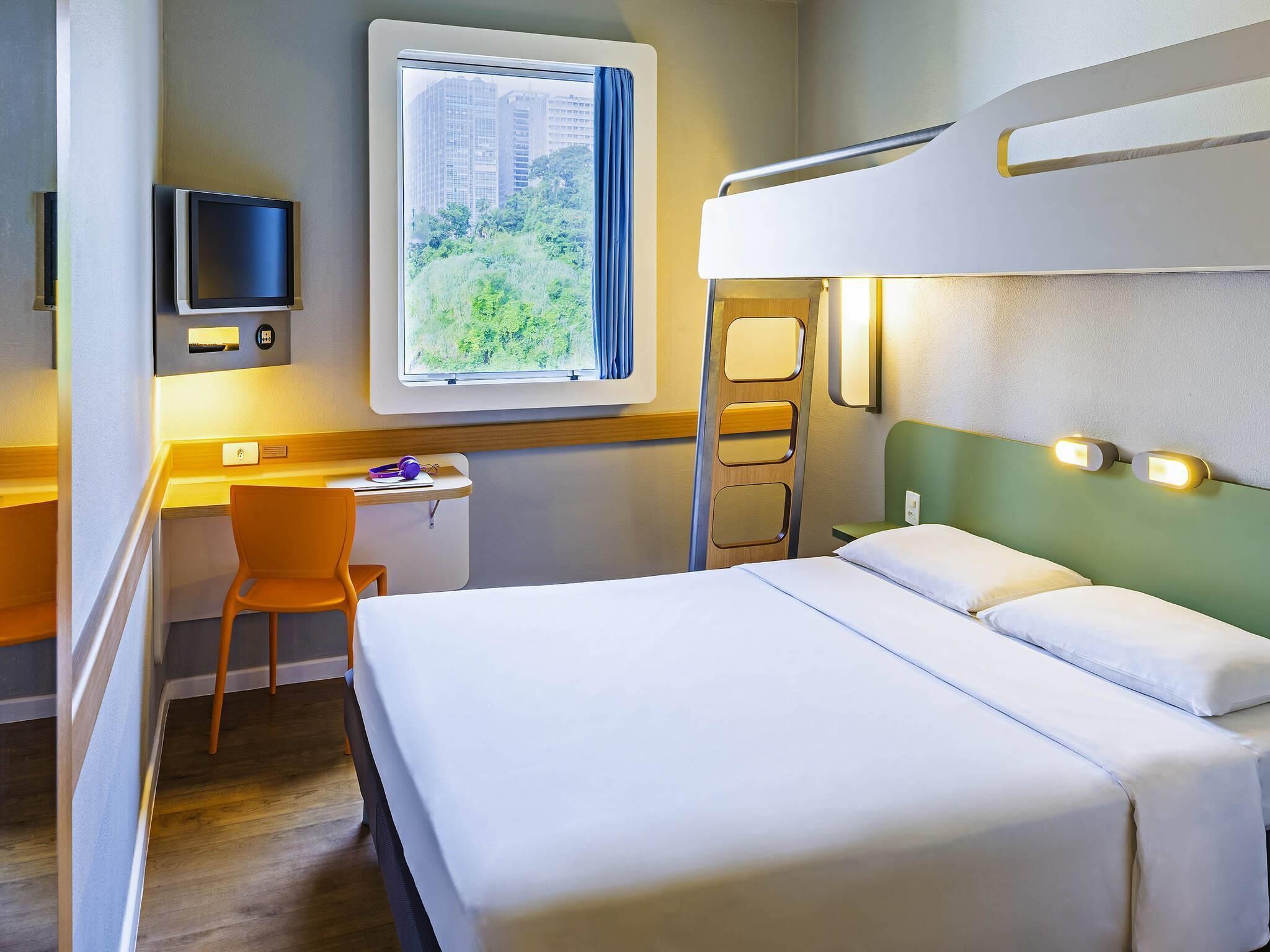 A hotel room with a bunk bed, a desk with a chair, a television, and a window with a view of buildings and greenery.