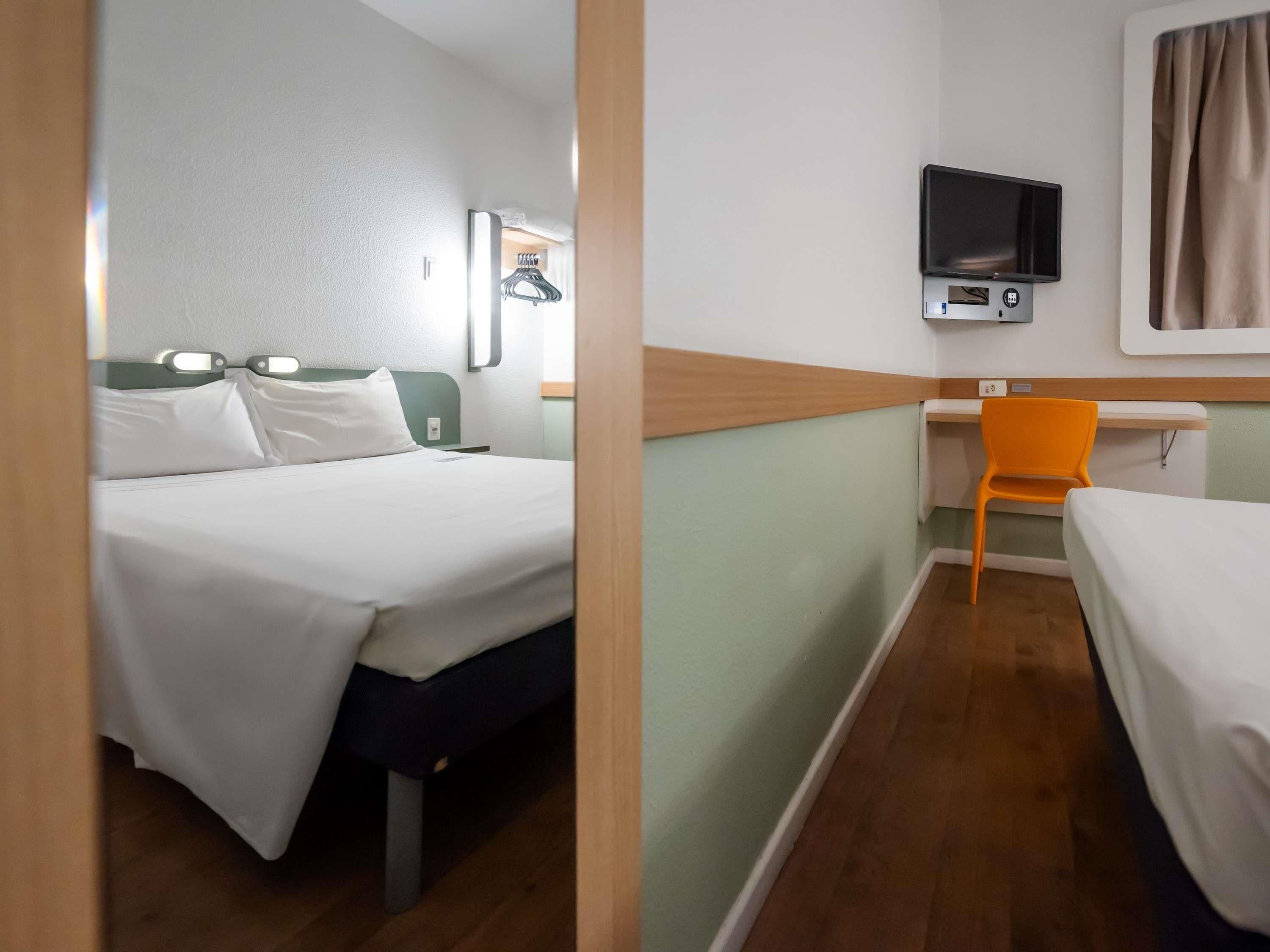 A hotel room with a bed, a desk with a chair, a television, and a window with curtains.