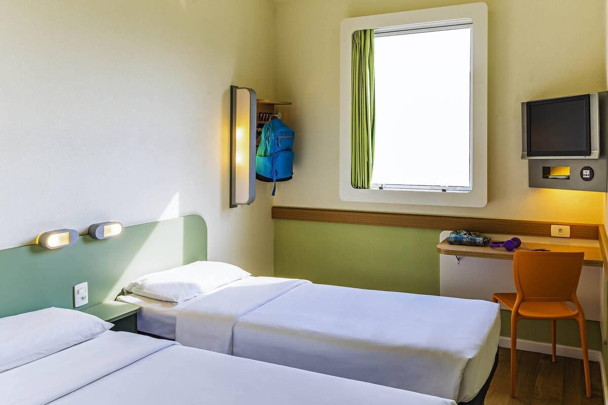 A hotel room with two beds, a desk with a chair, a TV, and a window with curtains.