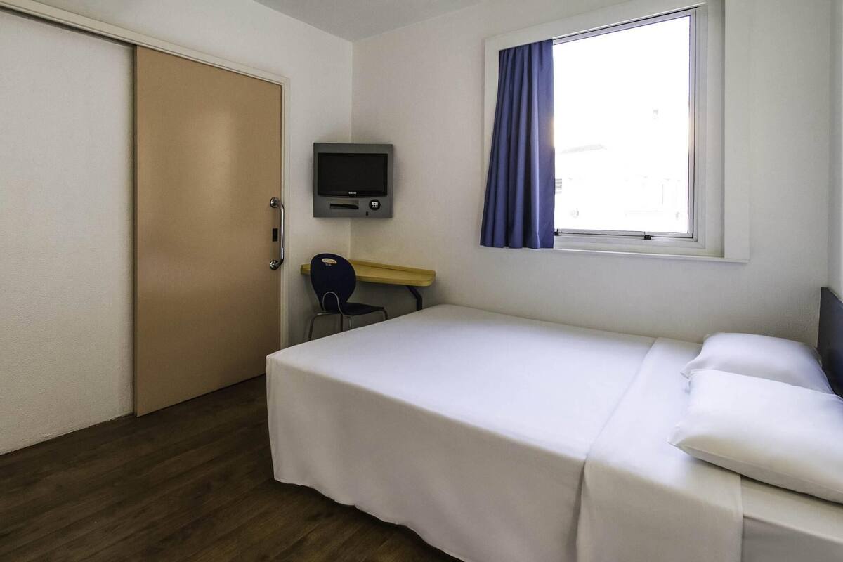 A hotel room with a bed, a desk, a chair, a television, and a window with curtains.