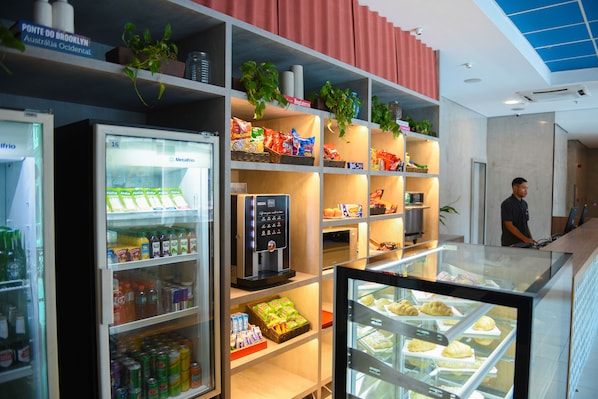 Um saguão de hotel com estação de bebidas de serviço próprio, prateleiras com lanches e uma vitrine de pastelaria.