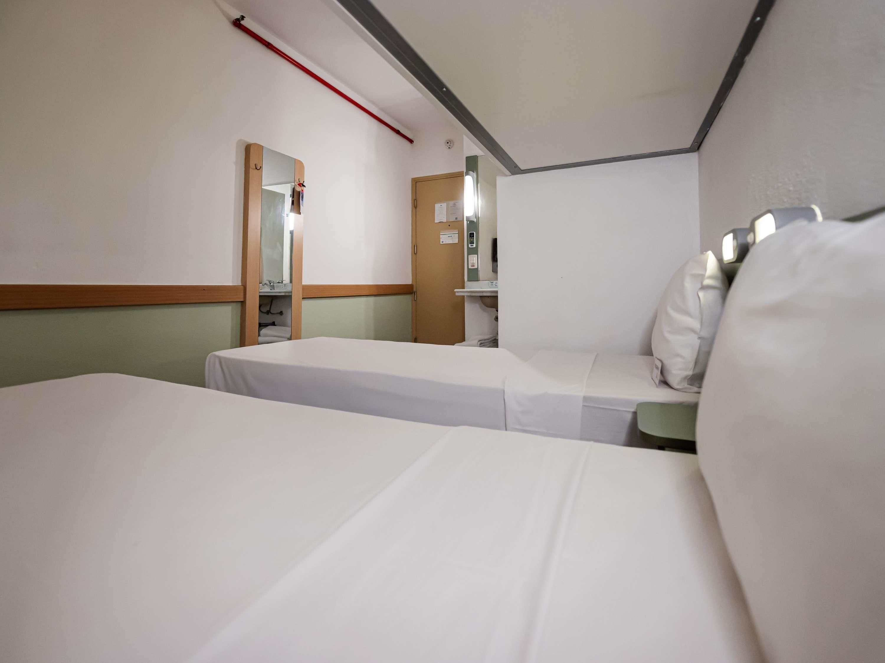 Quarto de hotel com duas camas, um espelho, mesa de cabeceira e janela.