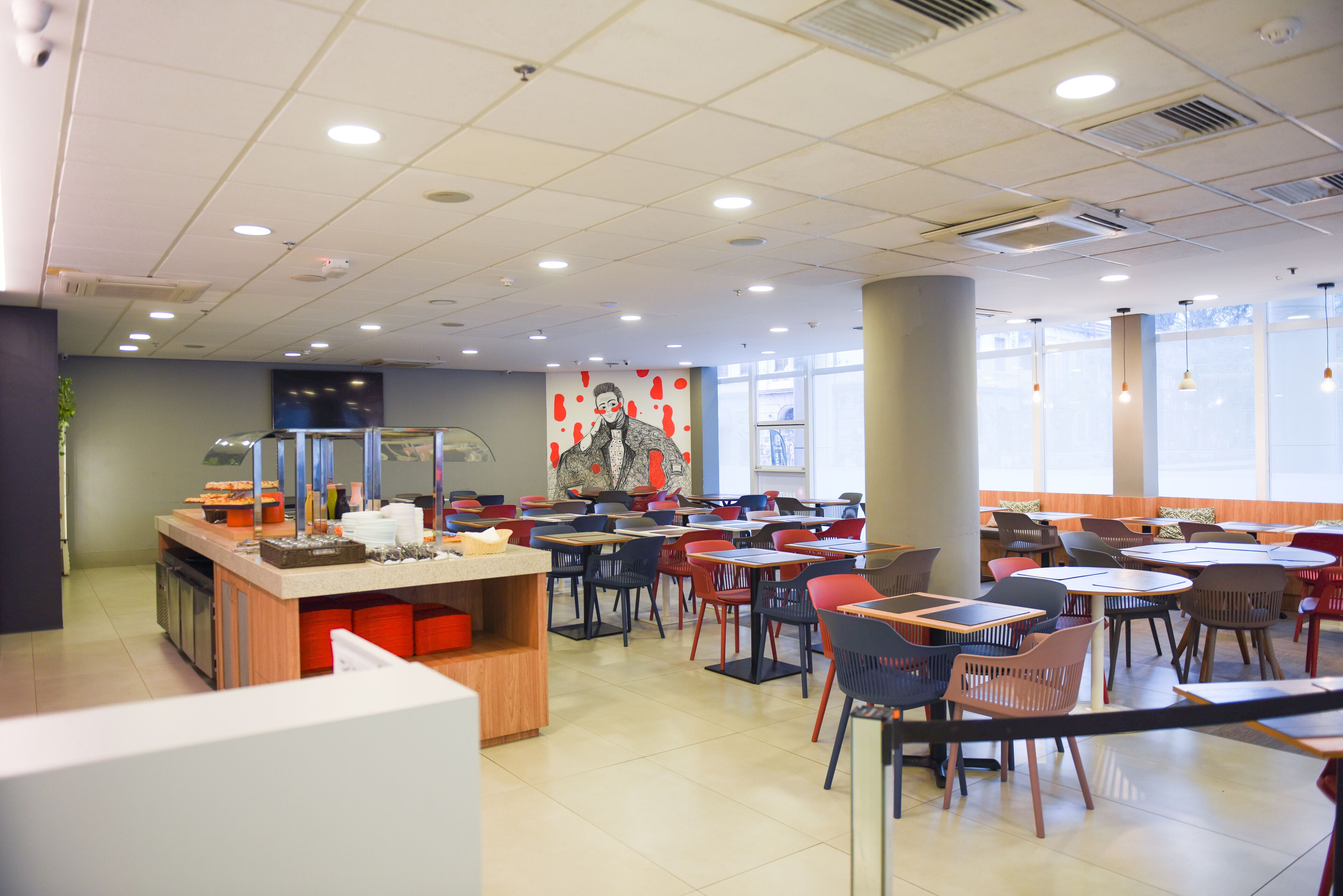 Una cafetería con varias mesas y sillas, una barra con alimentos y un amplio ventanal que proporciona luz natural.