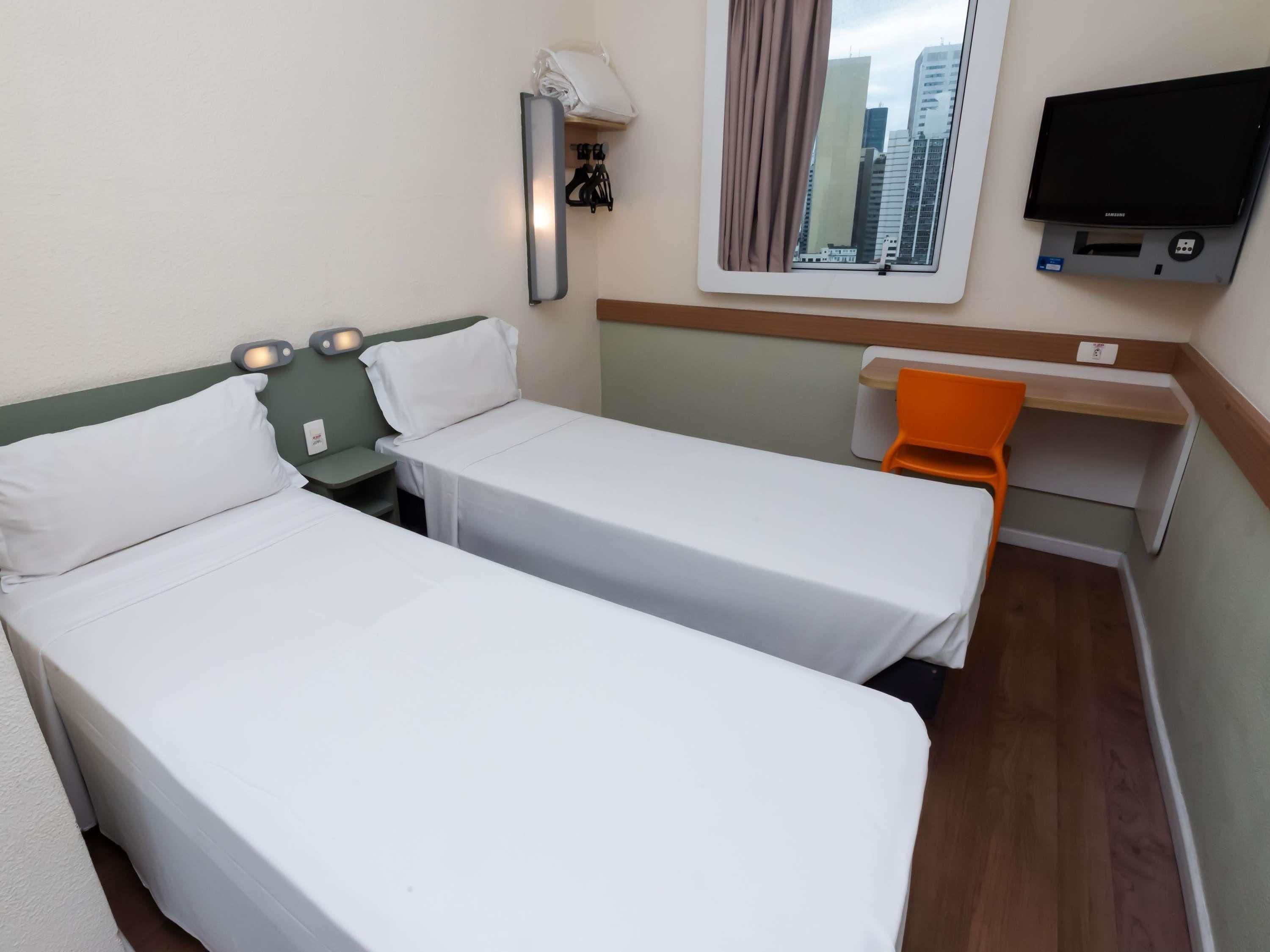 Une chambre d’hôtel avec deux lits, un bureau, une chaise orange, une télévision et une fenêtre donnant sur la ville.