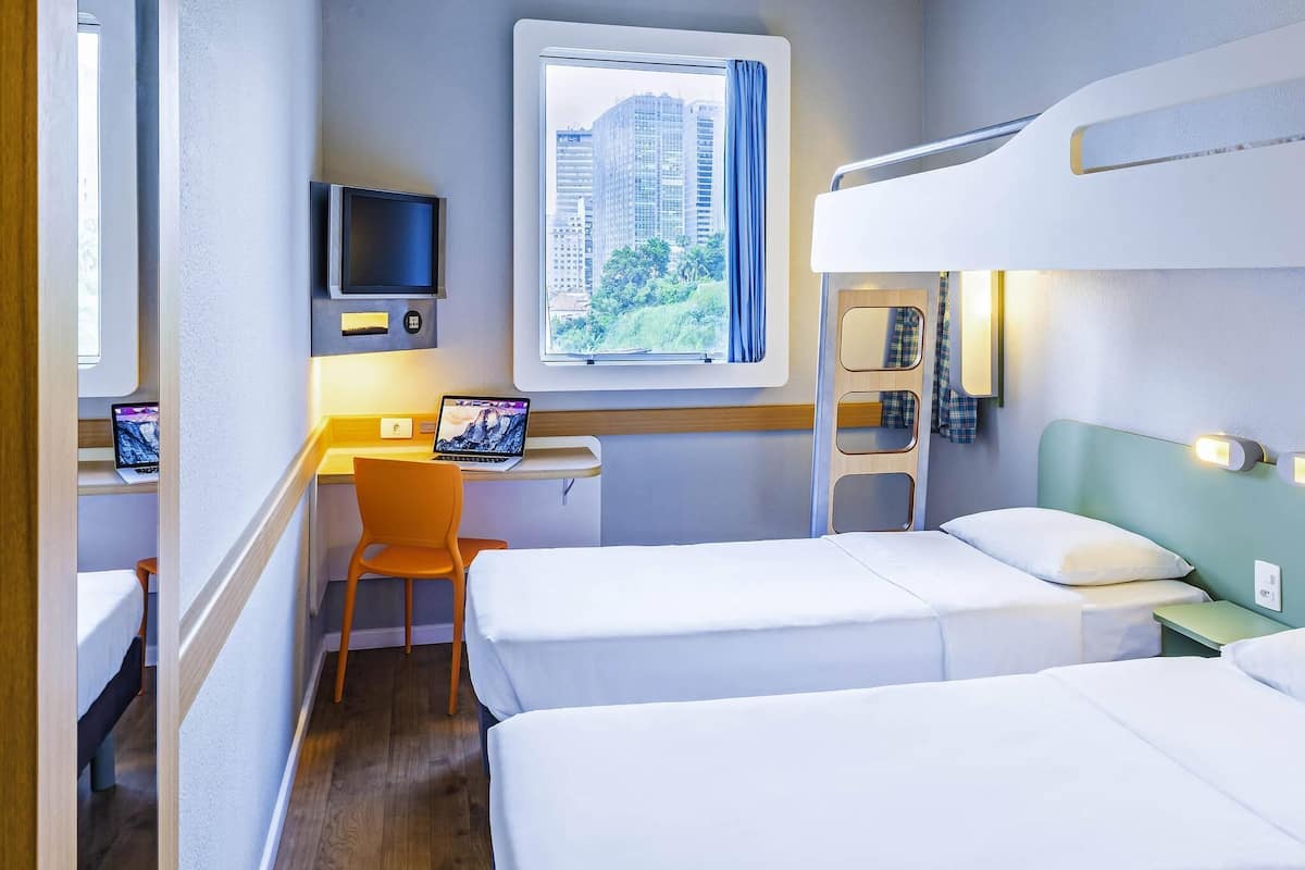 A hotel room with two beds, a desk with a laptop, a television, and a window with a city view.