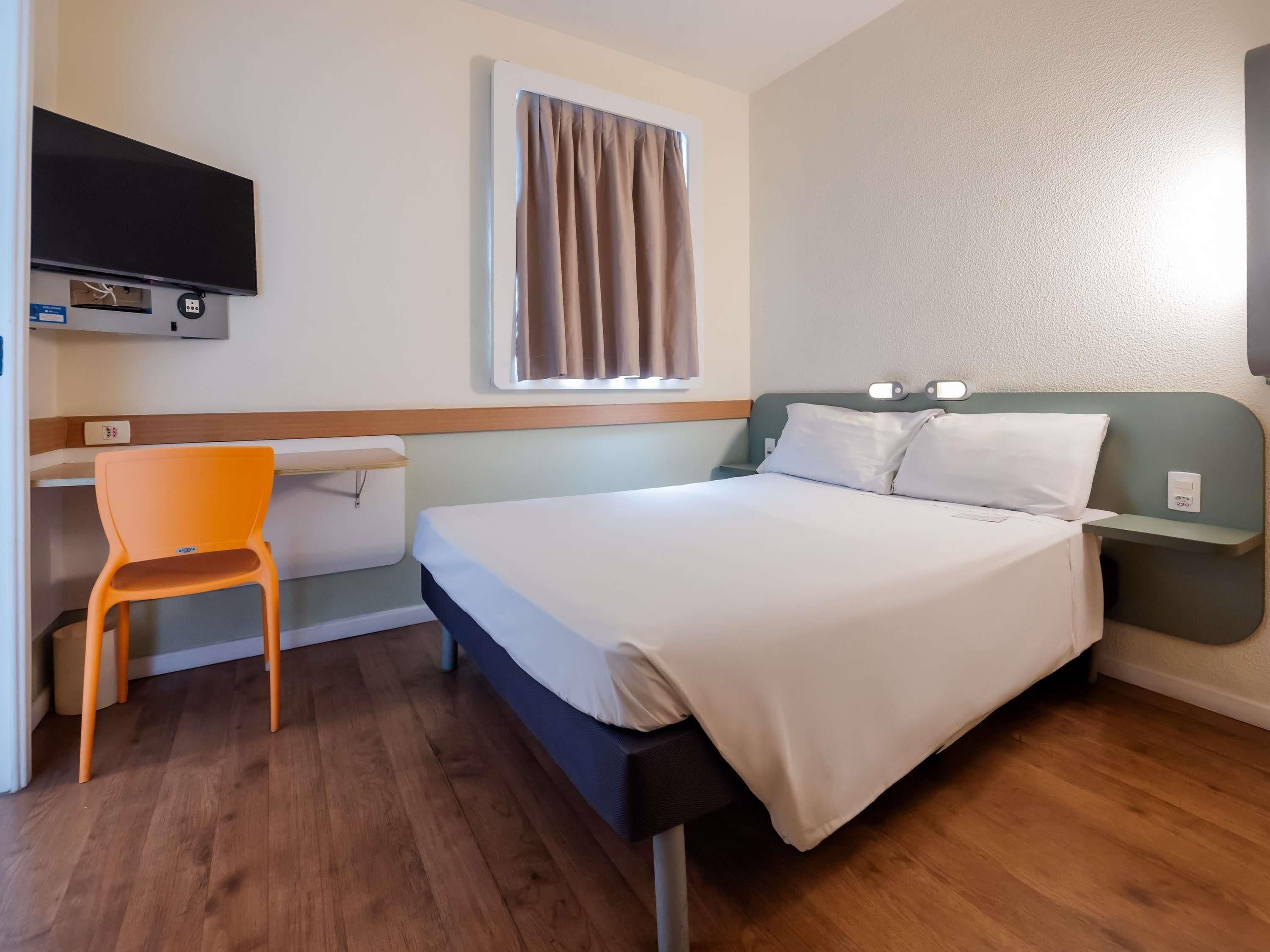 Habitación de hotel con cama, escritorio, silla, televisor y ventana con cortinas.