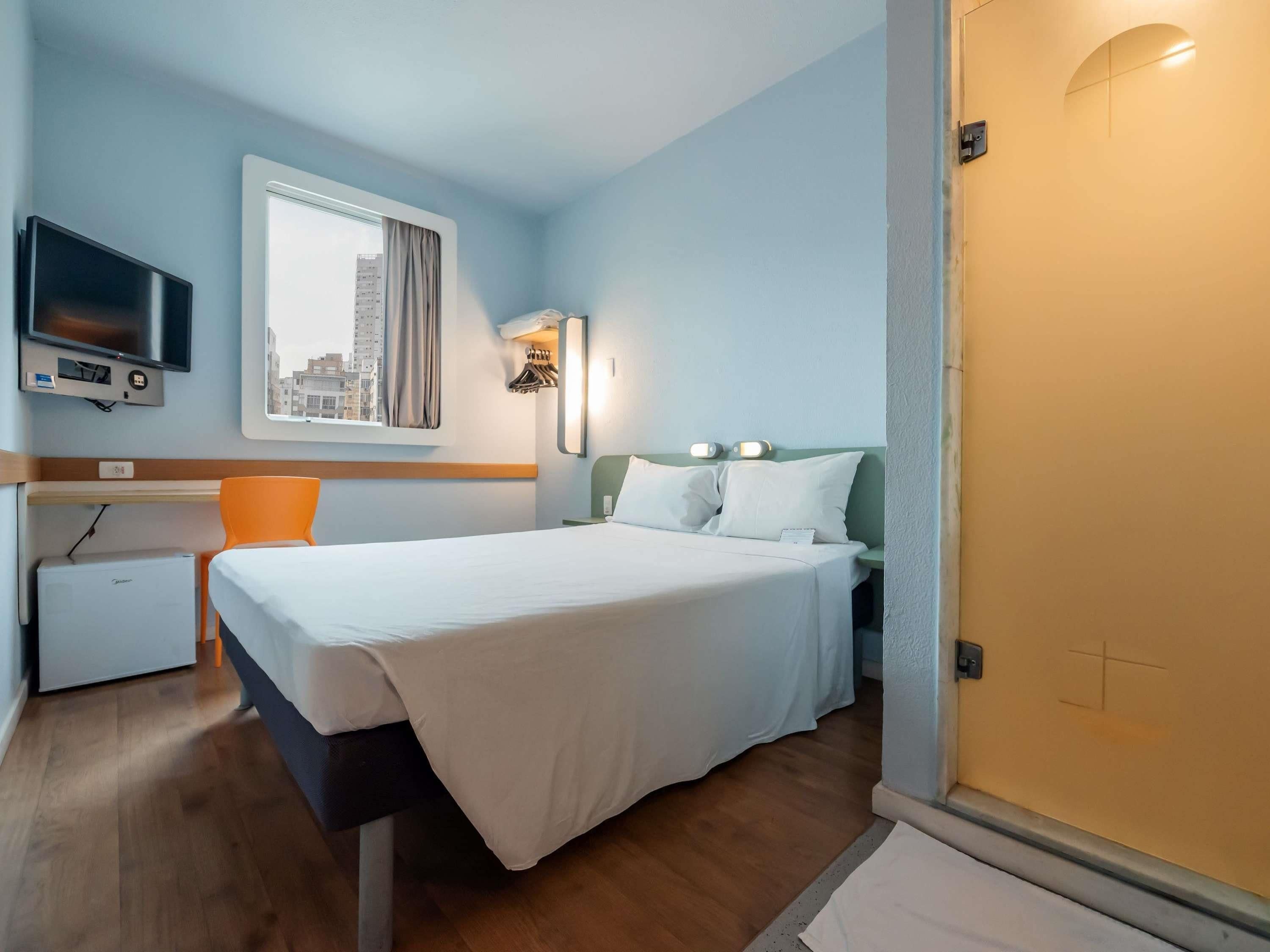 Quarto de hotel com cama, escrivaninha, televisão e janela com vista para prédios.