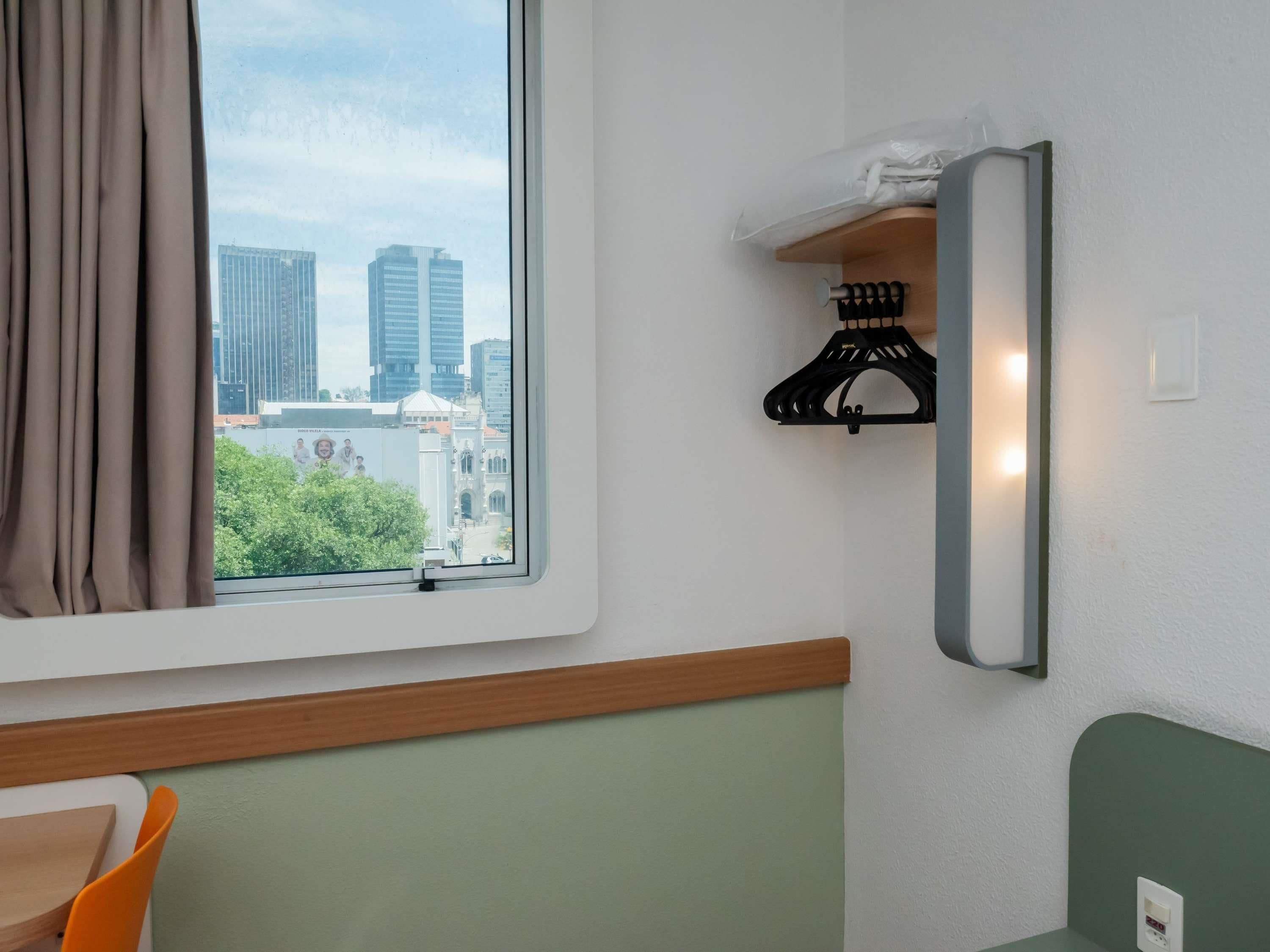 Ett hotellrum med ett fönster som vetter ut mot stadsbilden, en trähylla med en klädhängare och en grön vägg.