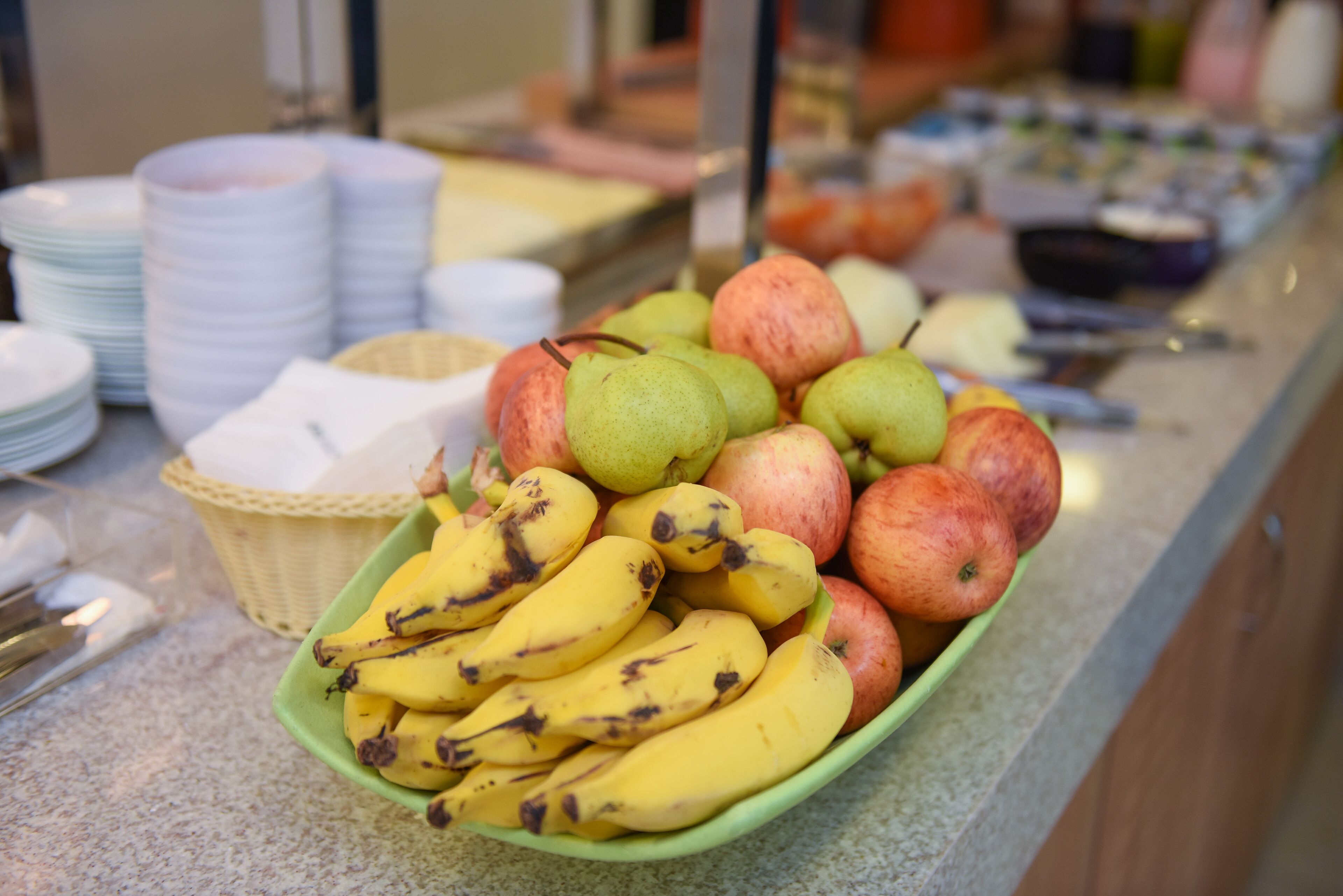 Mostrador de buffet con una variedad de frutas como plátanos, manzanas y peras, junto con platos blancos apilados y una cesta de mimbre.