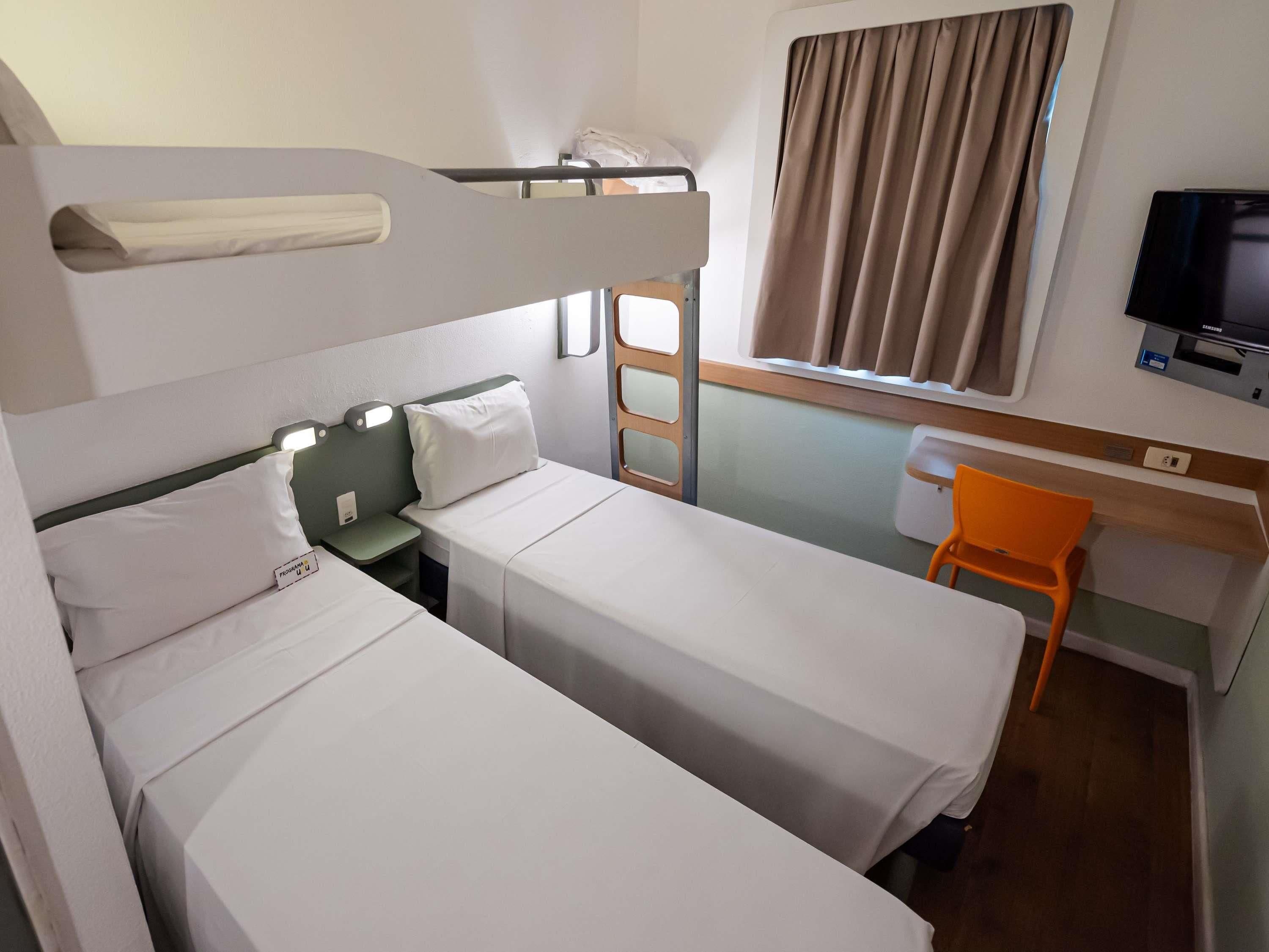 Een hotelkamer met twee enkele bedden, een bureau, een oranje stoel, een televisie en een raam met gordijnen.
