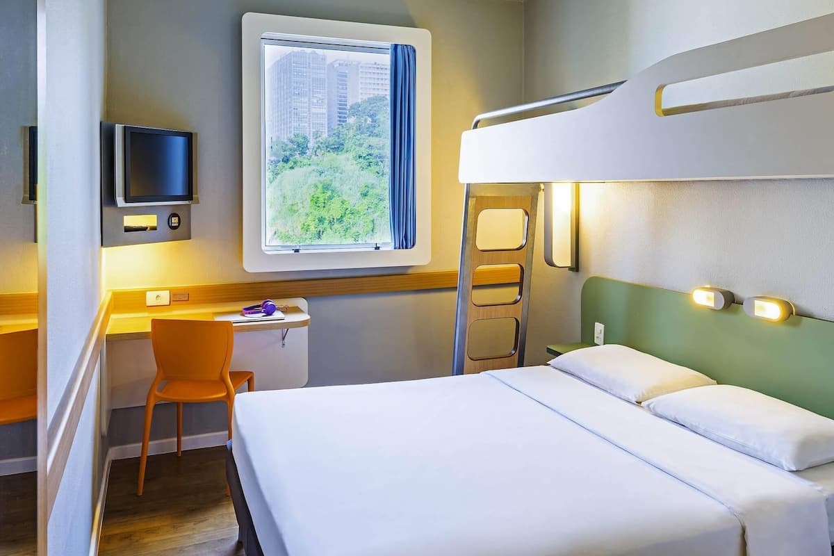 A hotel room with a bunk bed, a desk with a chair, a TV, and a window with a view of buildings and greenery.