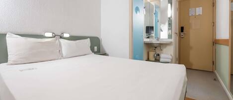 A hotel room with a bed, a bedside table, a mirror, a bathroom with a sink, and a door.