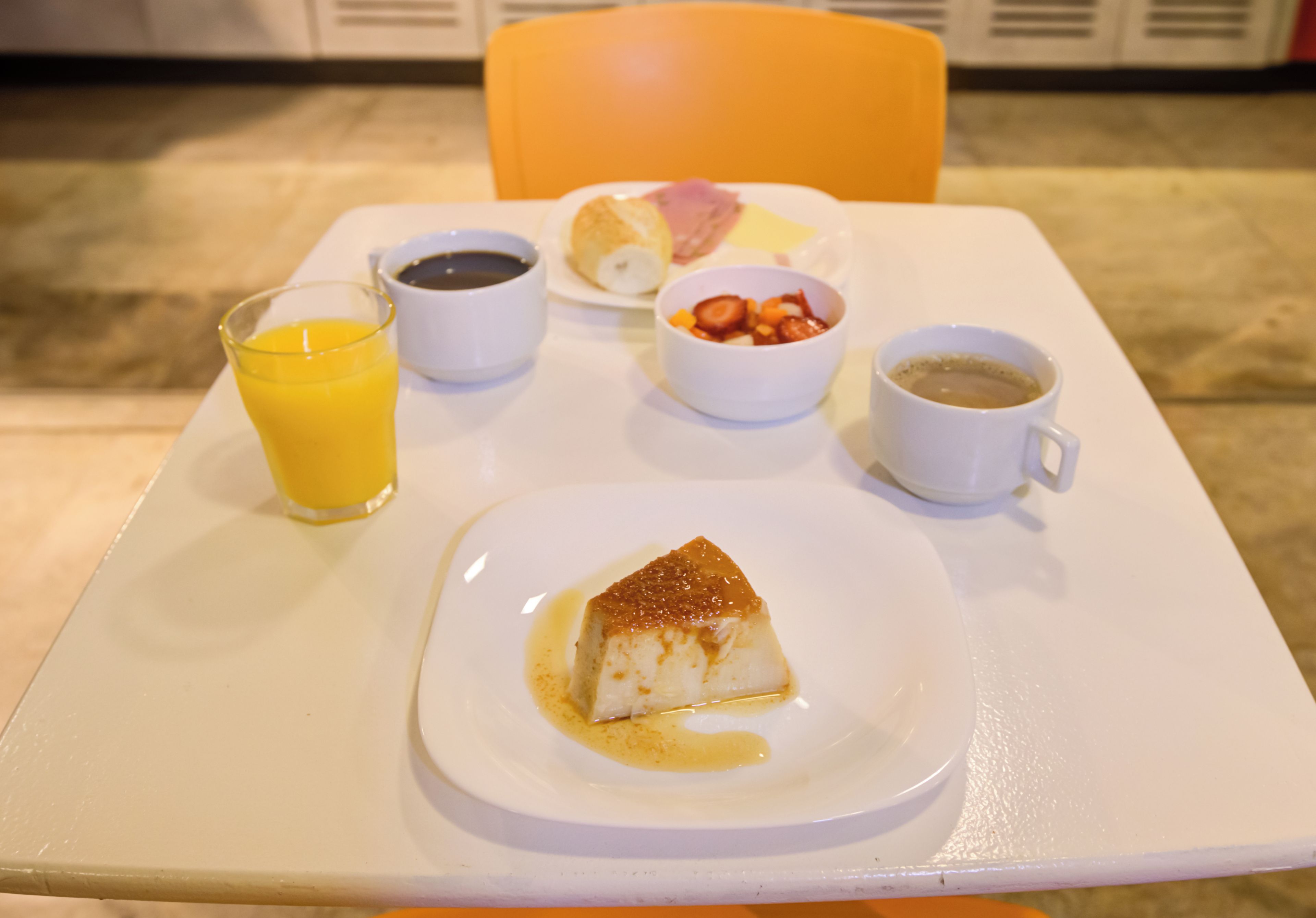 A breakfast tray with a slice of flan, a glass of orange juice, a cup of coffee, a bowl of fruit, and a roll on a white table.