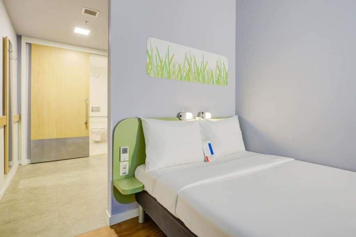 A hotel room with a bed, a green chair, a wall-mounted light, and a door leading to another room.