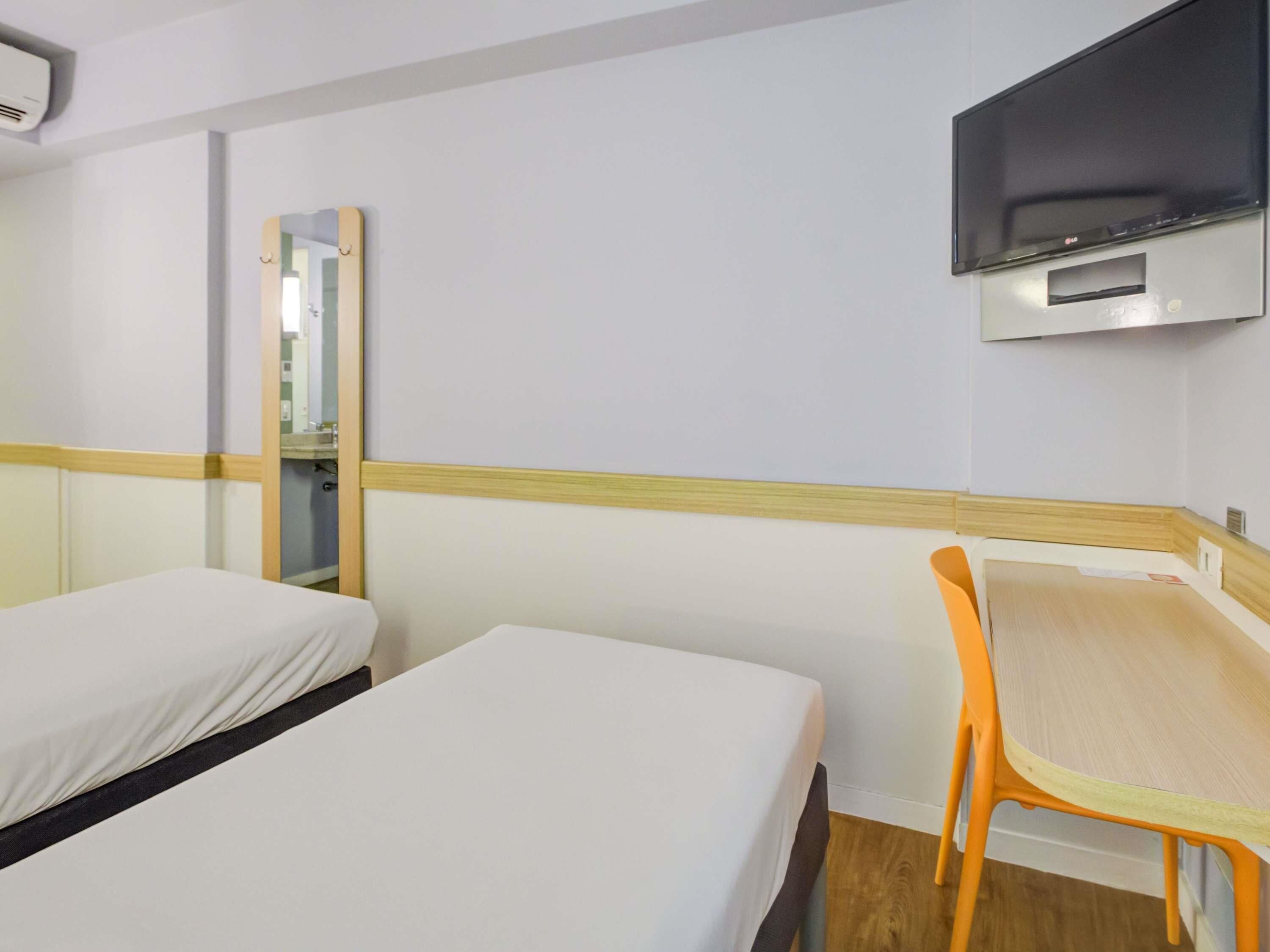 Ein Hotelzimmer mit zwei Betten, einem Schreibtisch, einem Sessel, einem Fernseher und einem Spiegel.
