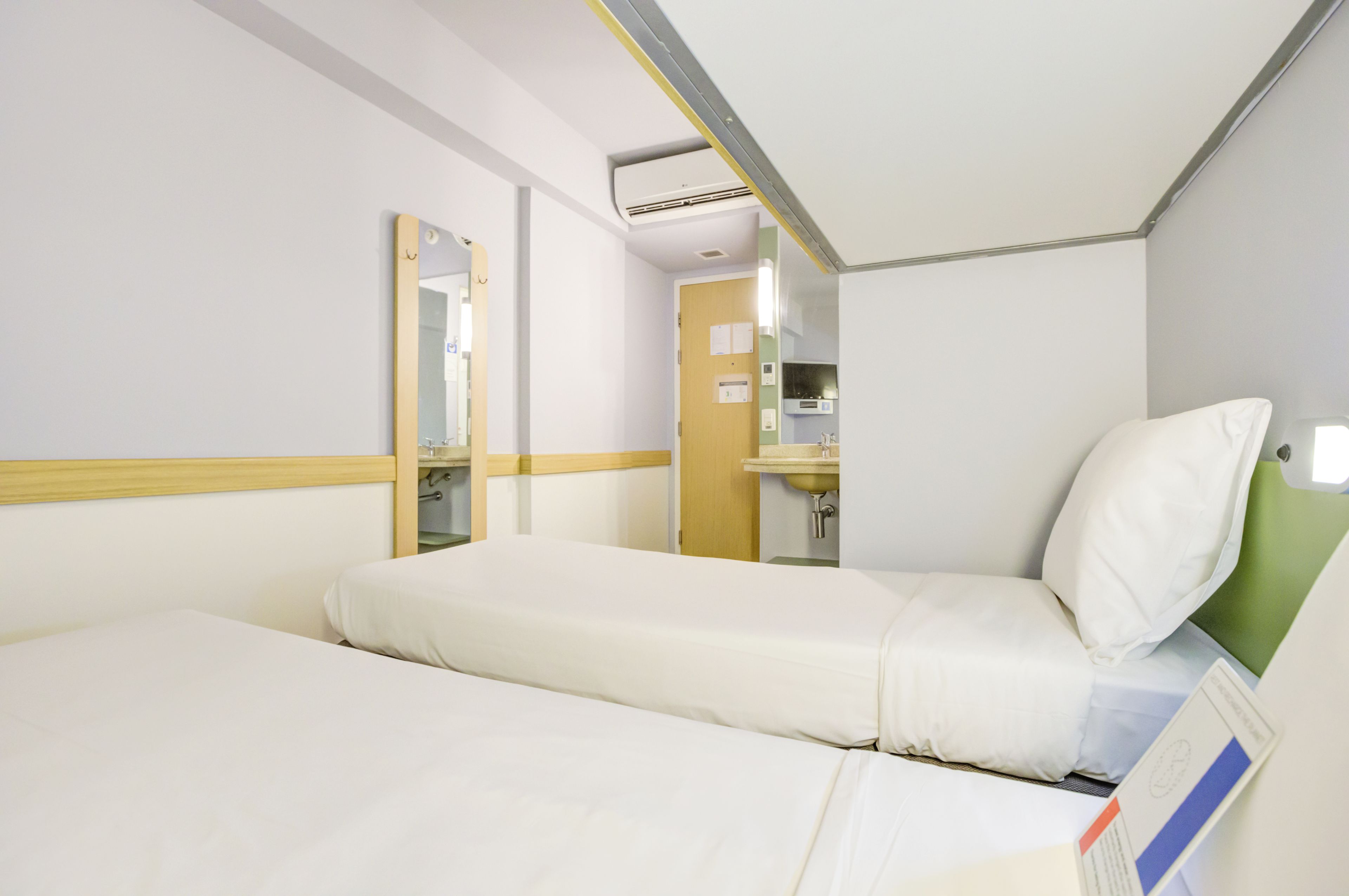 Habitación de hotel con una cama individual, una mesita de noche, un espejo, un ventanal pequeño y un equipo de aire acondicionado instalado en la pared.