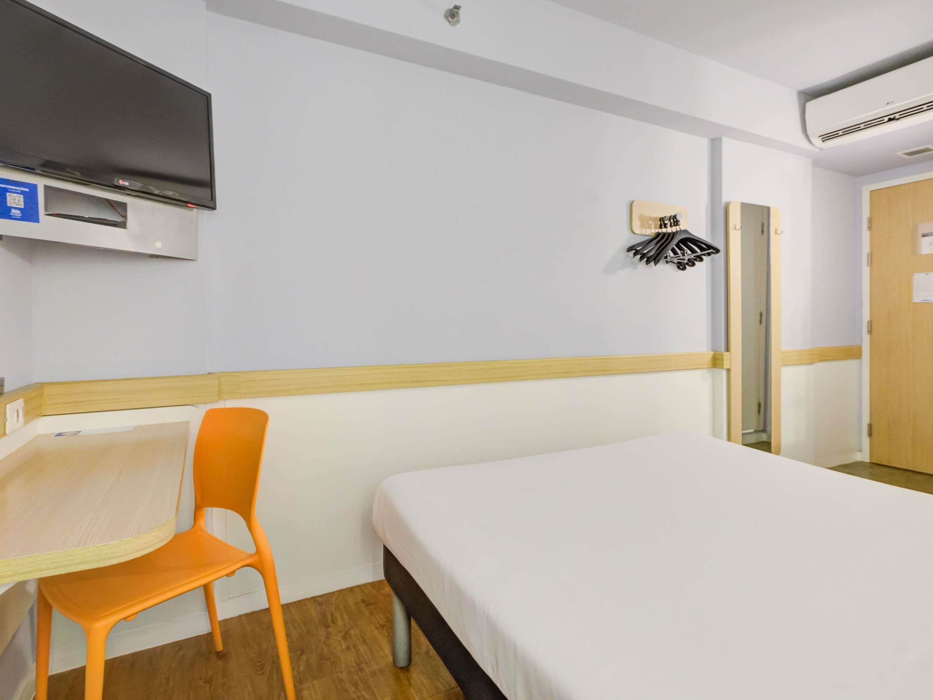 Una habitación de hotel con cama, escritorio, una silla naranja, televisor de pantalla plana y toallero de pared.