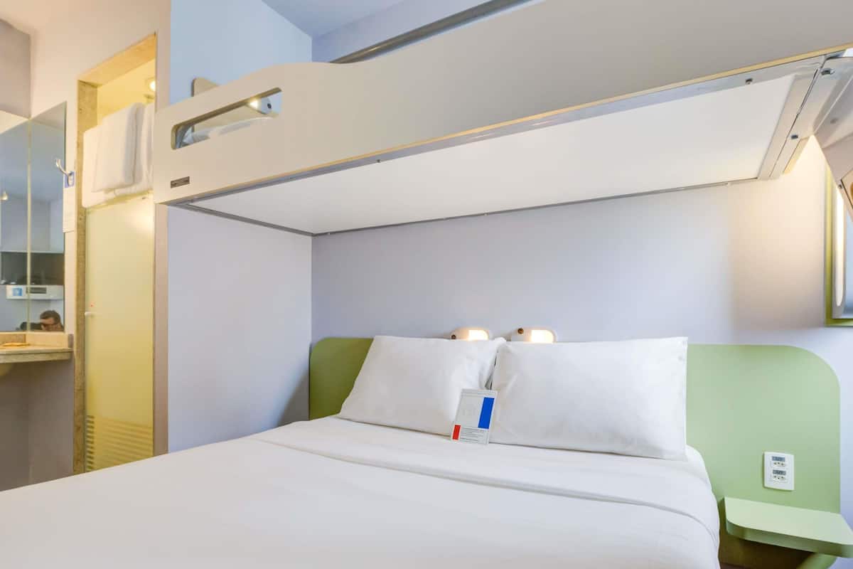 A hotel room with a bunk bed, a bedside table, a nightstand, a mirror, and a window.