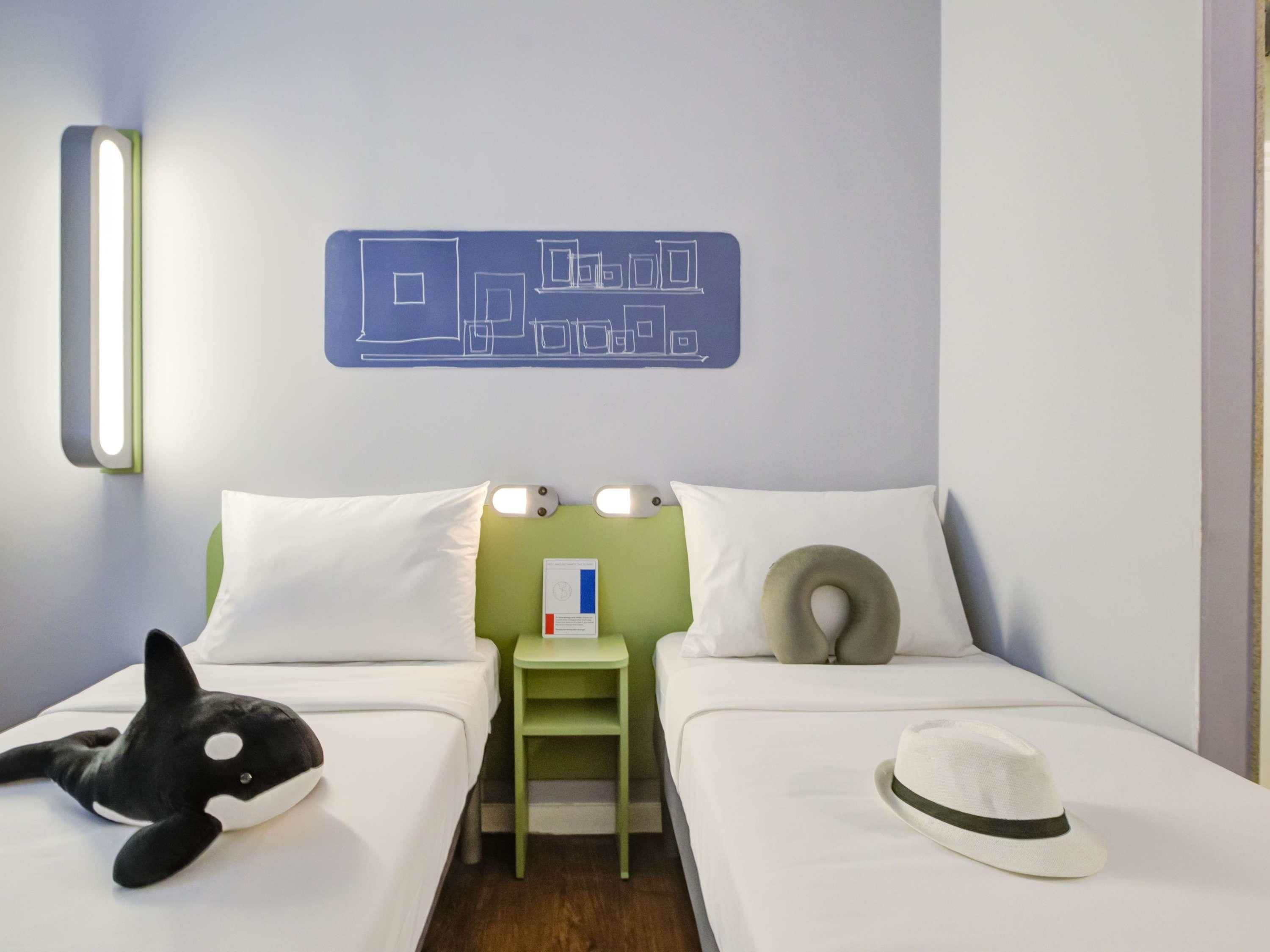 Quarto de hotel com duas camas, uma mesa de cabeceira verde, um brinquedo de orca macio, um chapéu branco e um luminário fixado na parede.