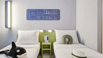 Quarto de hotel com duas camas, uma mesa de cabeceira verde, um brinquedo de orca macio, um chapéu branco e um luminário fixado na parede.