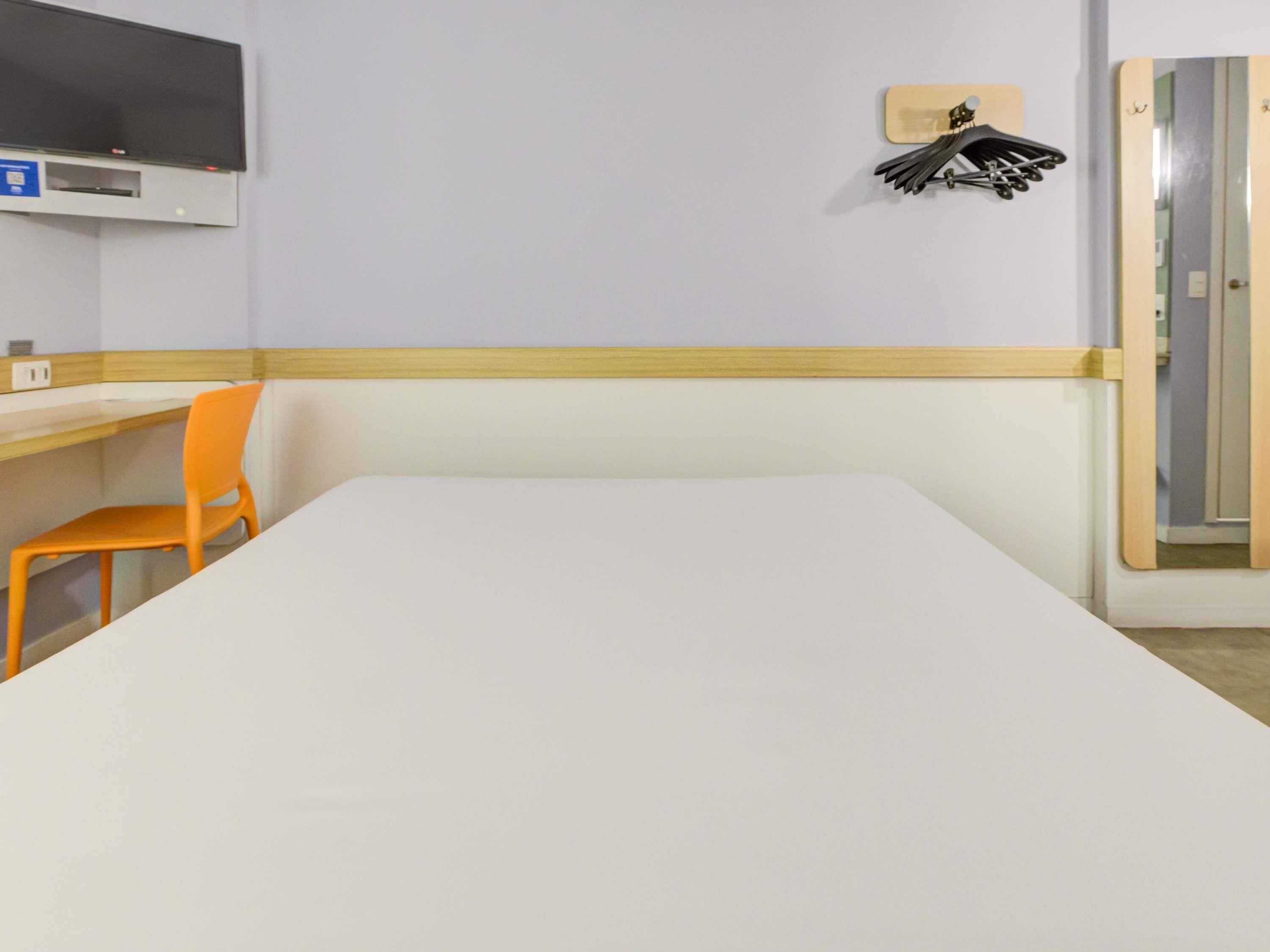 Quarto de hotel com uma cama grande, uma cadeira laranja, uma televisão na parede e um cabideiro com chapéus.