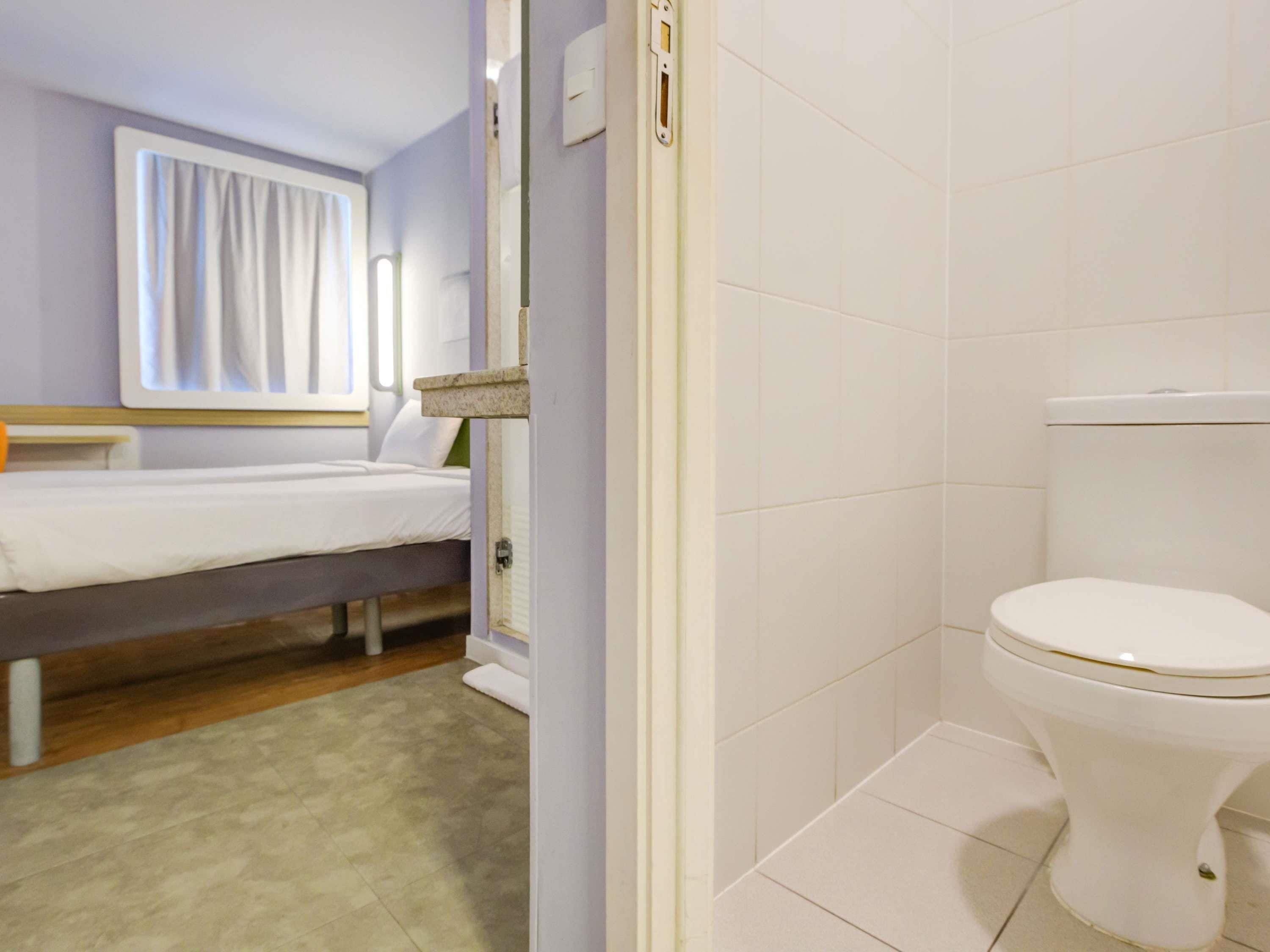 A bedroom with a bed, a bedside table, a window with curtains, and a bathroom with a toilet.