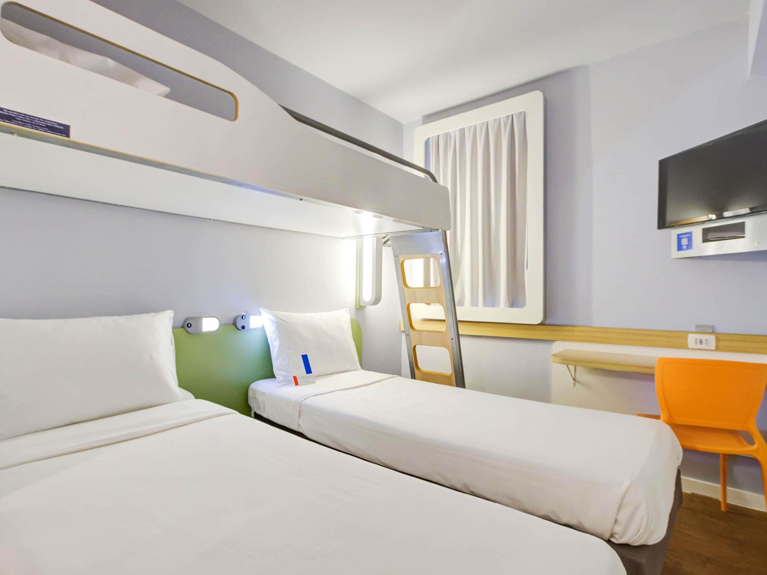 Quarto de hotel com duas camas, uma escrivaninha, uma cadeira, uma televisão e uma janela com cortinas.