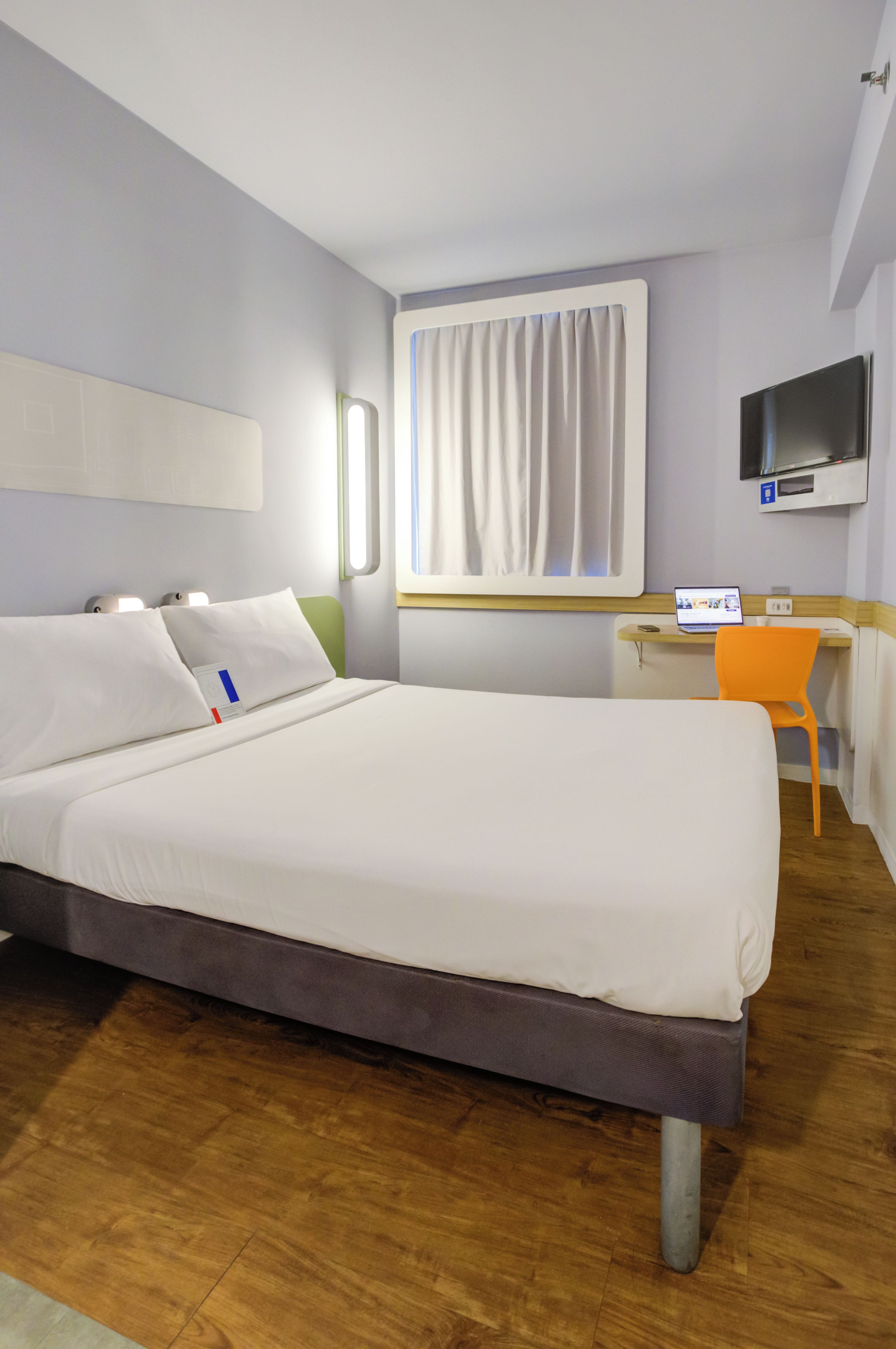 Habitació d'hotel amb llit, escriptori, cadira, televisió i finestra amb cortines.