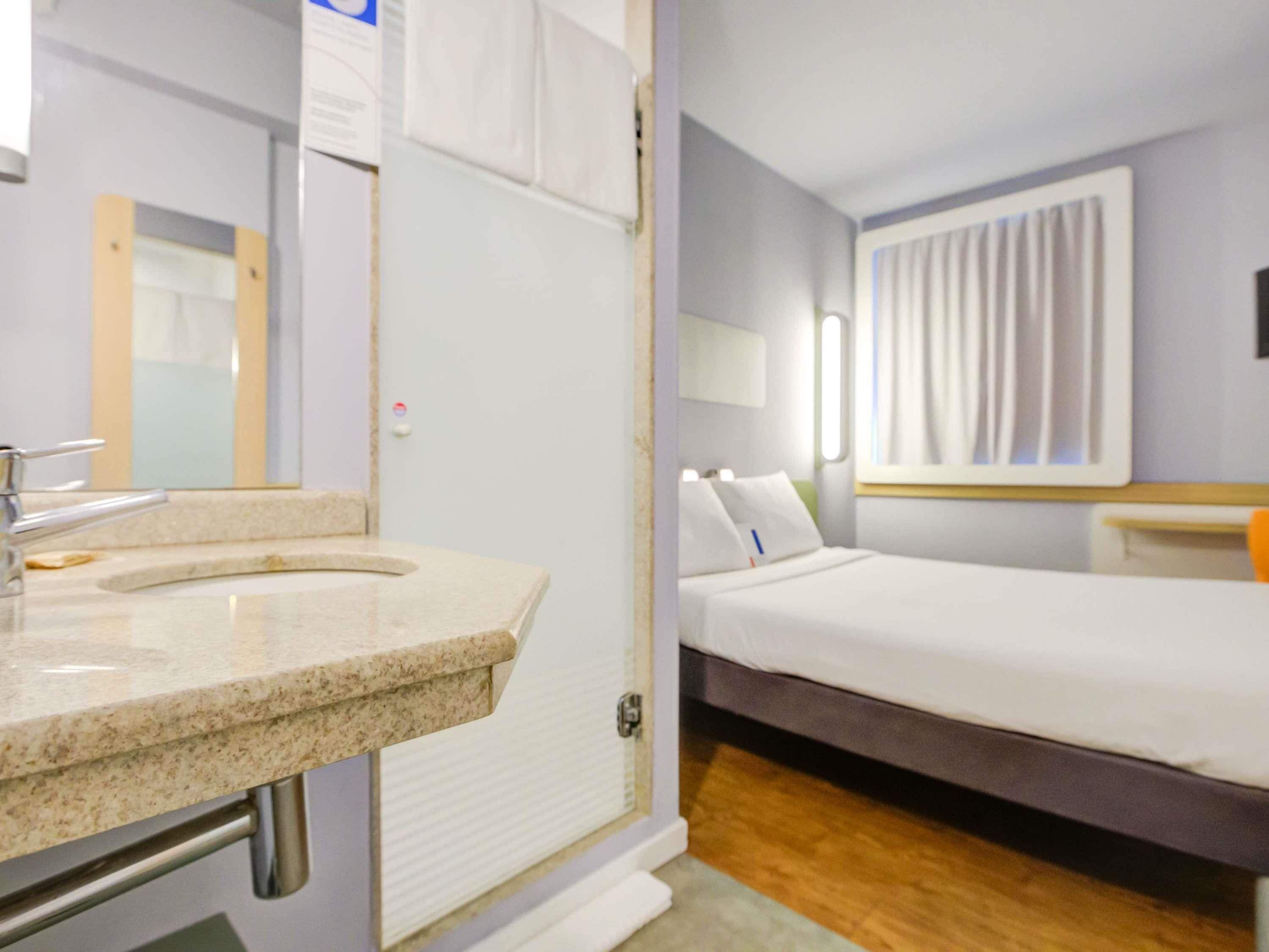 A hotel room with a bathroom, a bed, a nightstand, and a window with curtains.