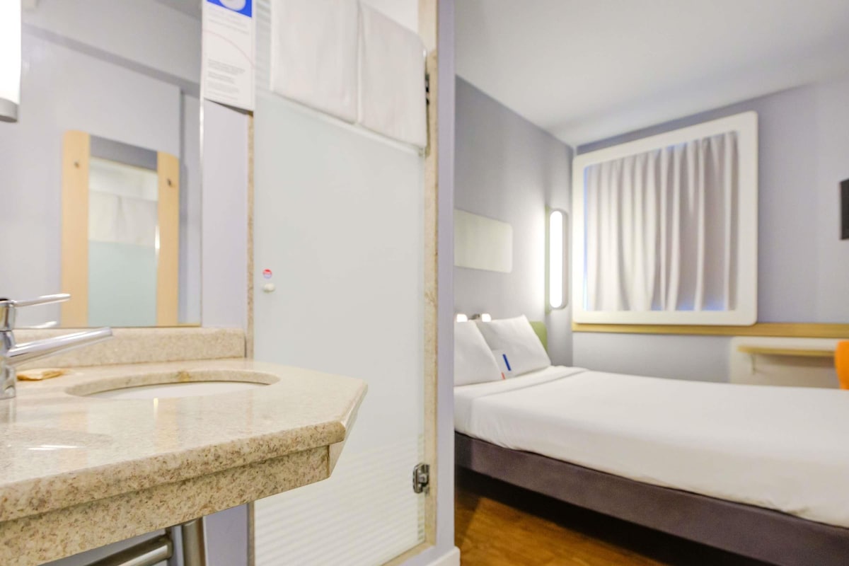 A hotel room with a bathroom, a bed, a nightstand, and a window with curtains.