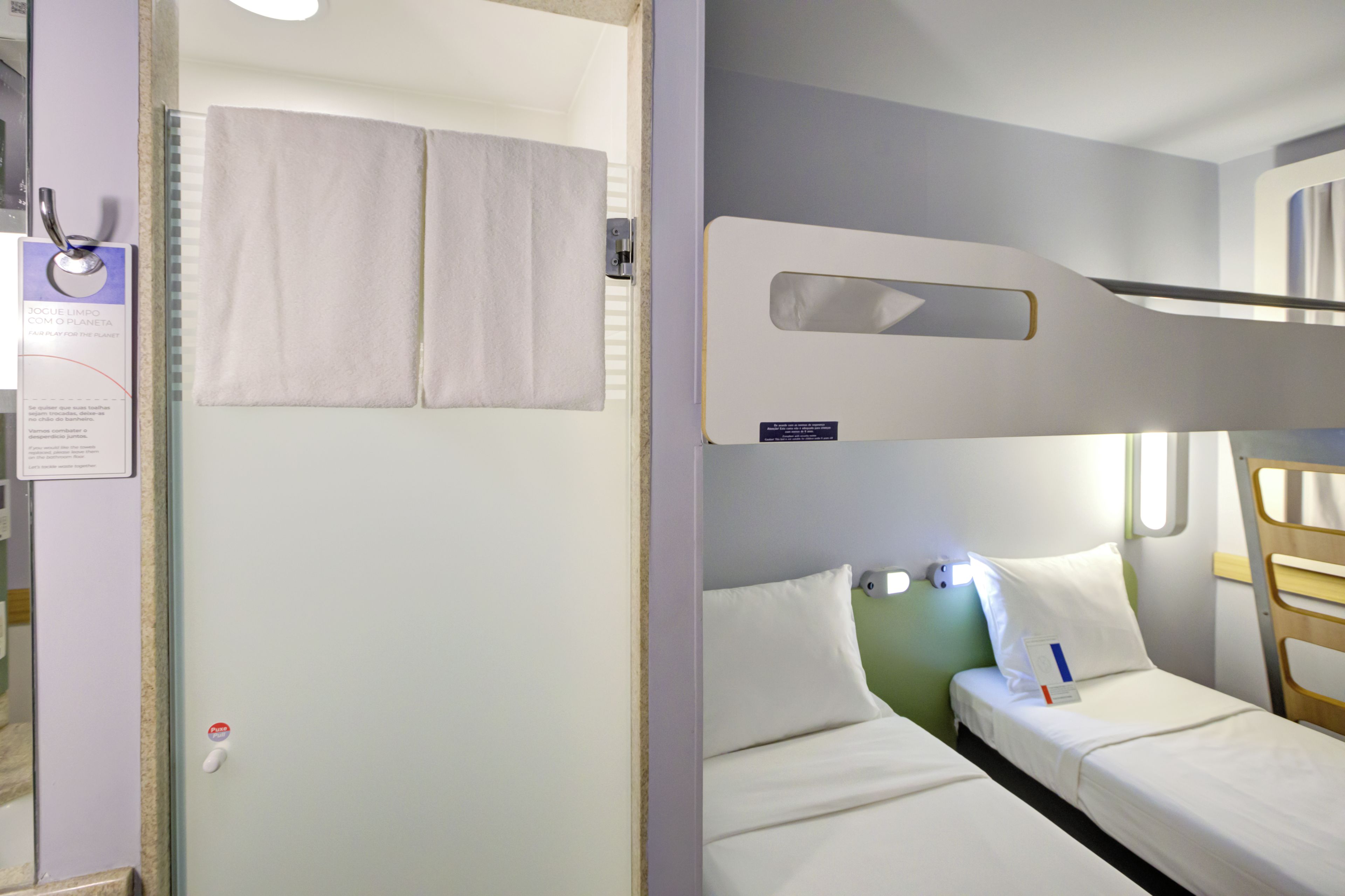 Une chambre d’hôtel avec un lit superposé, un lit simple, une salle de bain avec un porte-serviettes et une poignée de porte visible.