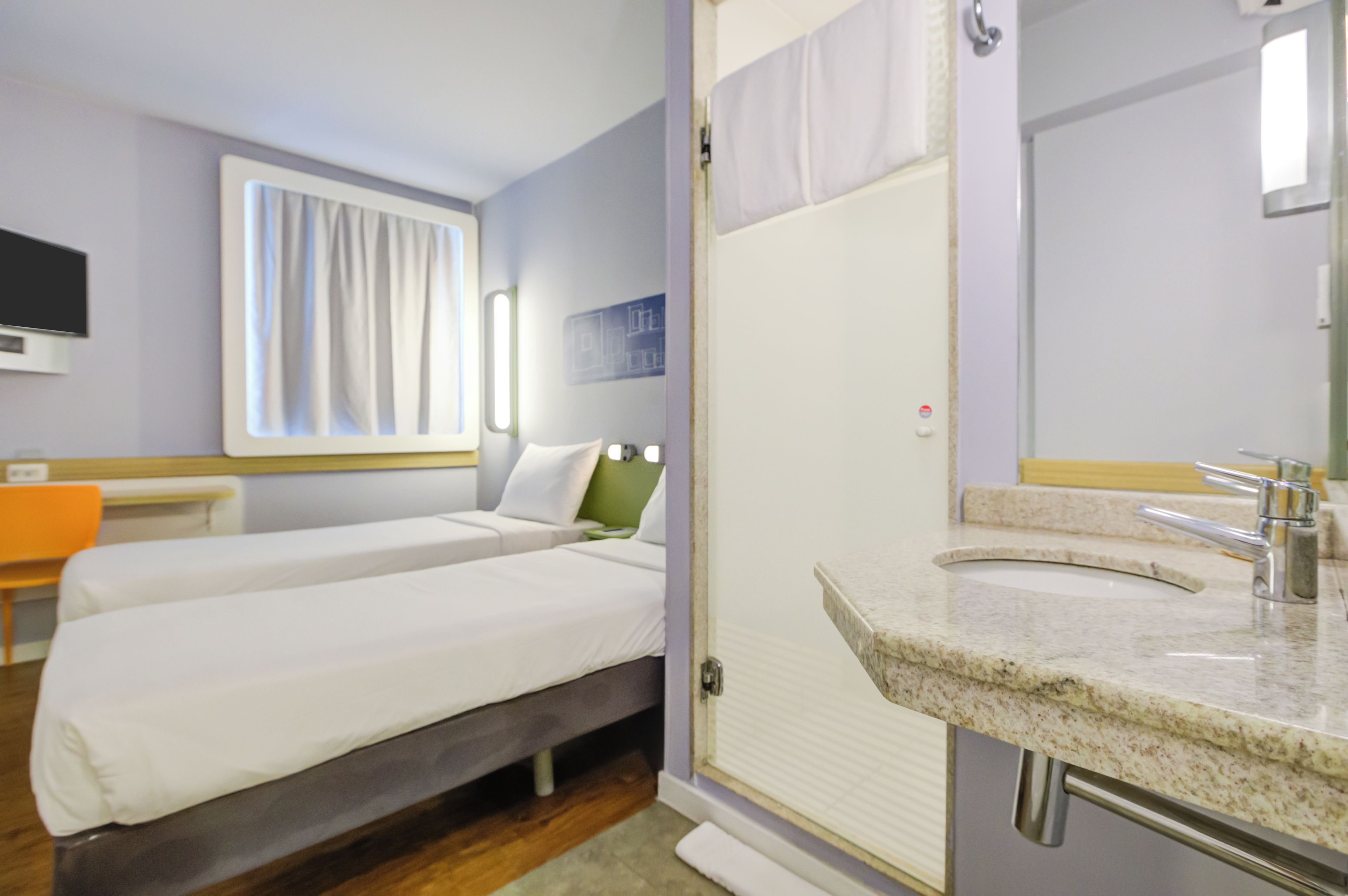 Habitación de hotel con dos camas, baño con lavamanos y espejo, y televisión en la pared.