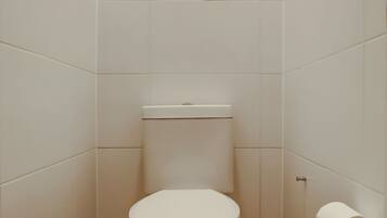 A small, white-tiled bathroom with a toilet, a toilet paper holder, and a flush handle.
