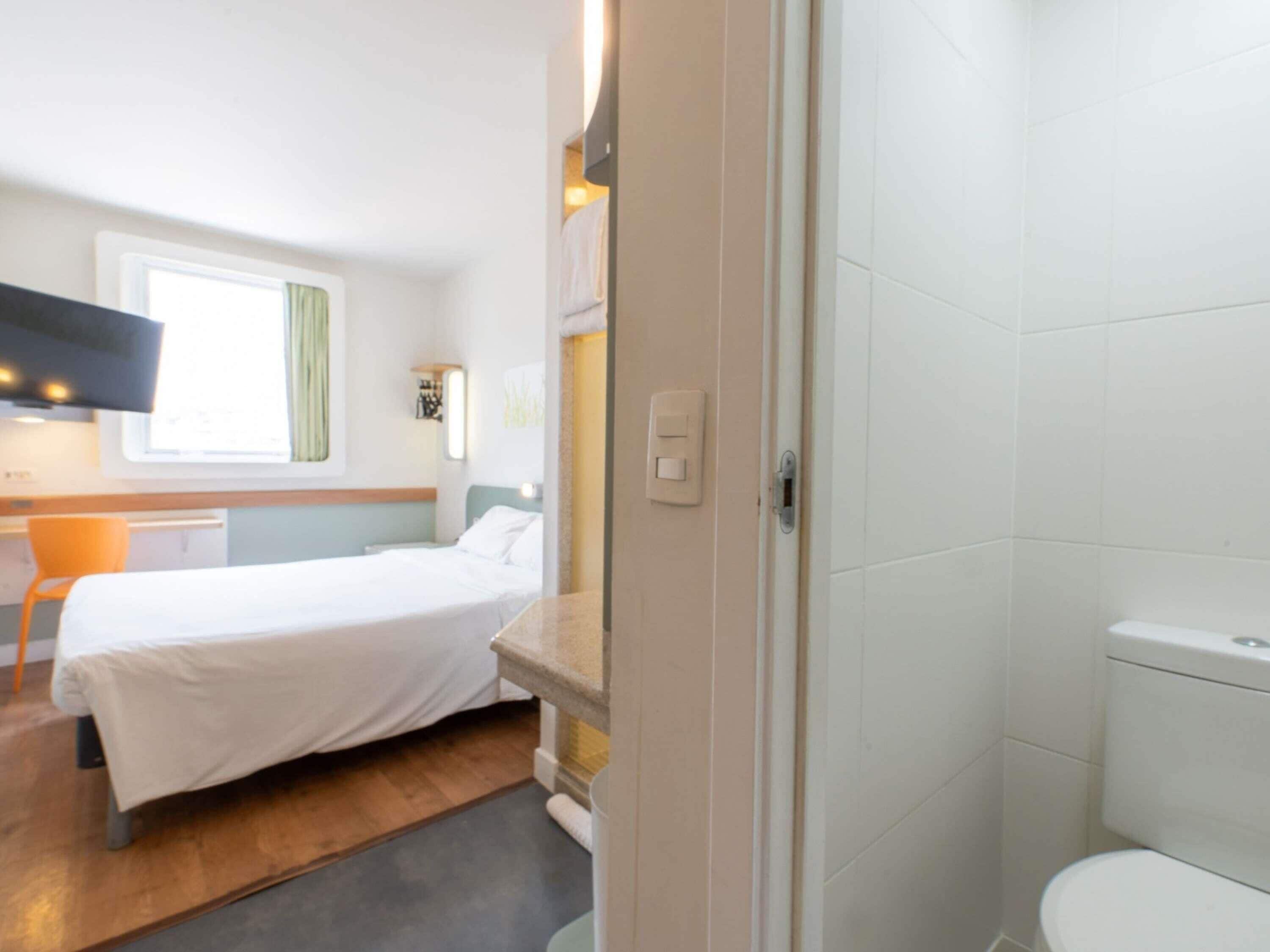 A compact hotel room with a bed, a small desk, a chair, a toilet, and a shower area.
