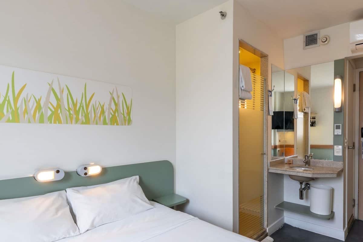 A hotel room with a bed, a wall-mounted light, a bathroom with a sink and mirror, and a door leading to another room.