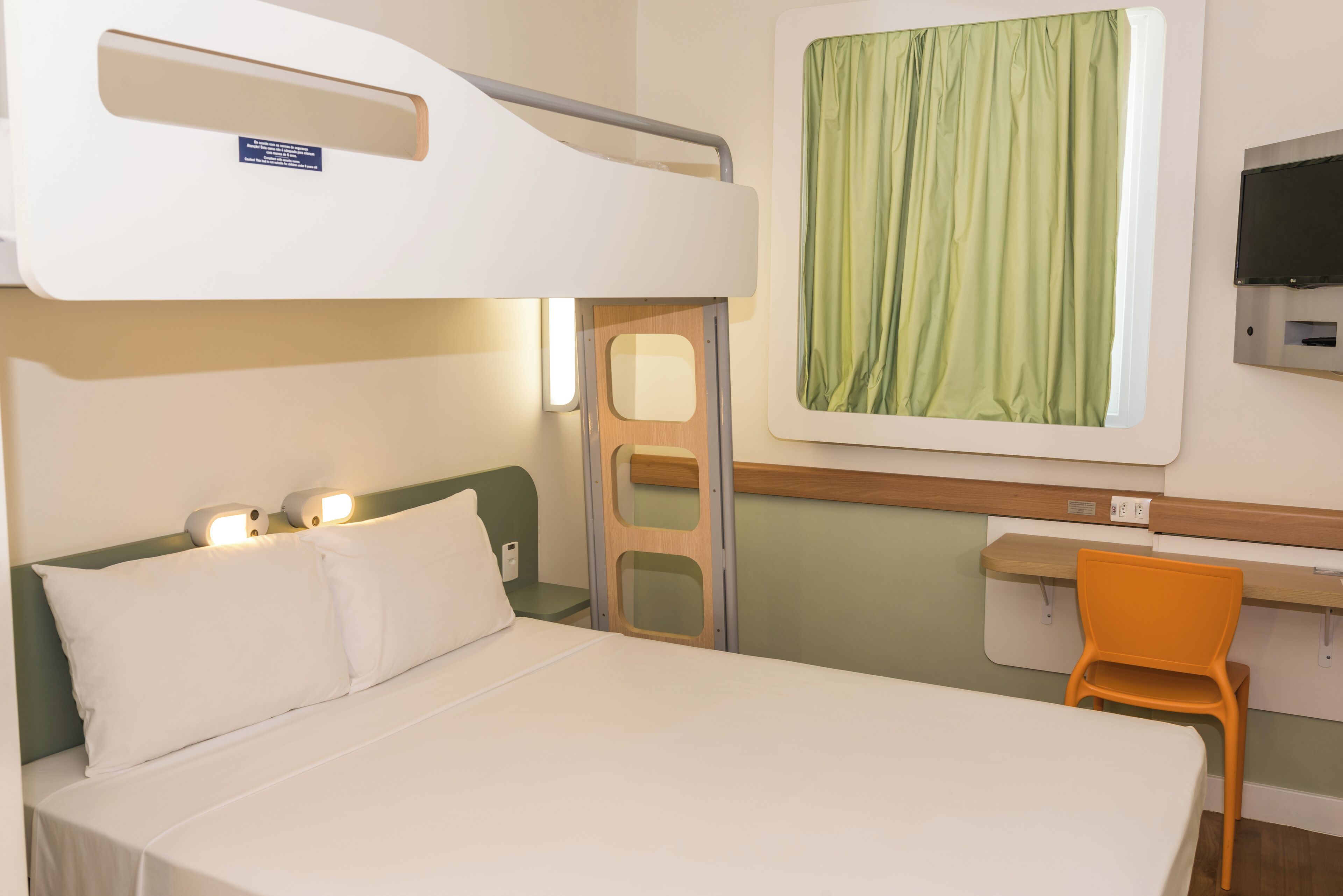 Una cama en literas con cabecera, un escritorio con silla, un televisor y una ventana con cortinas.