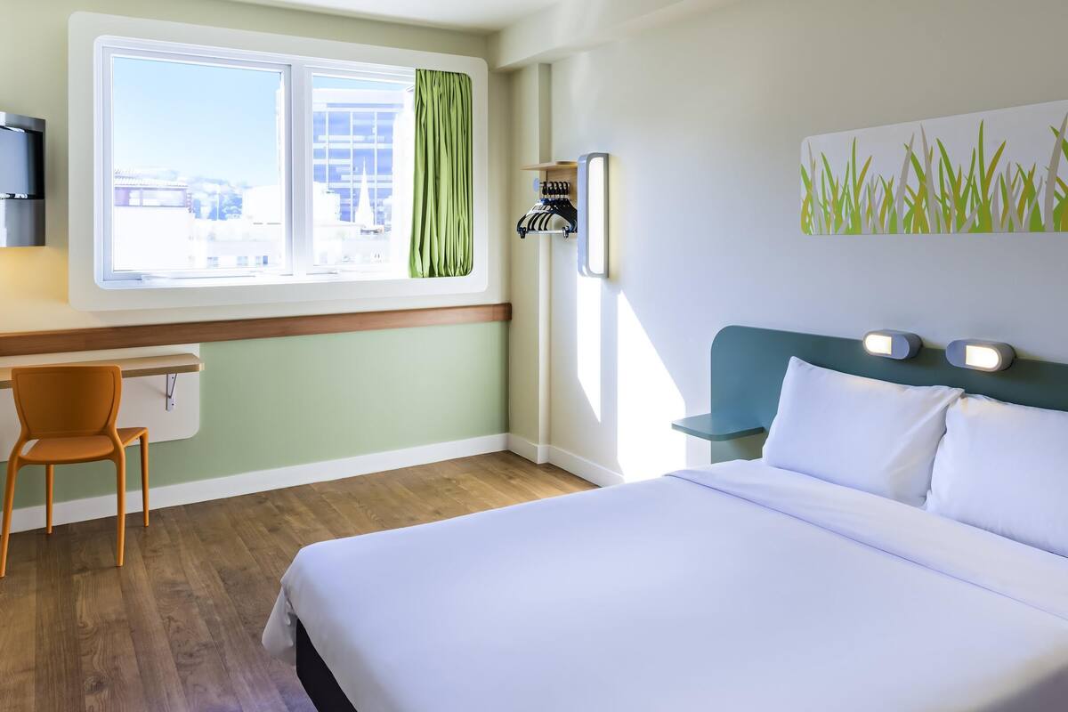 A hotel room with a bed, a desk, a chair, a window with curtains, and a wall-mounted light fixture.