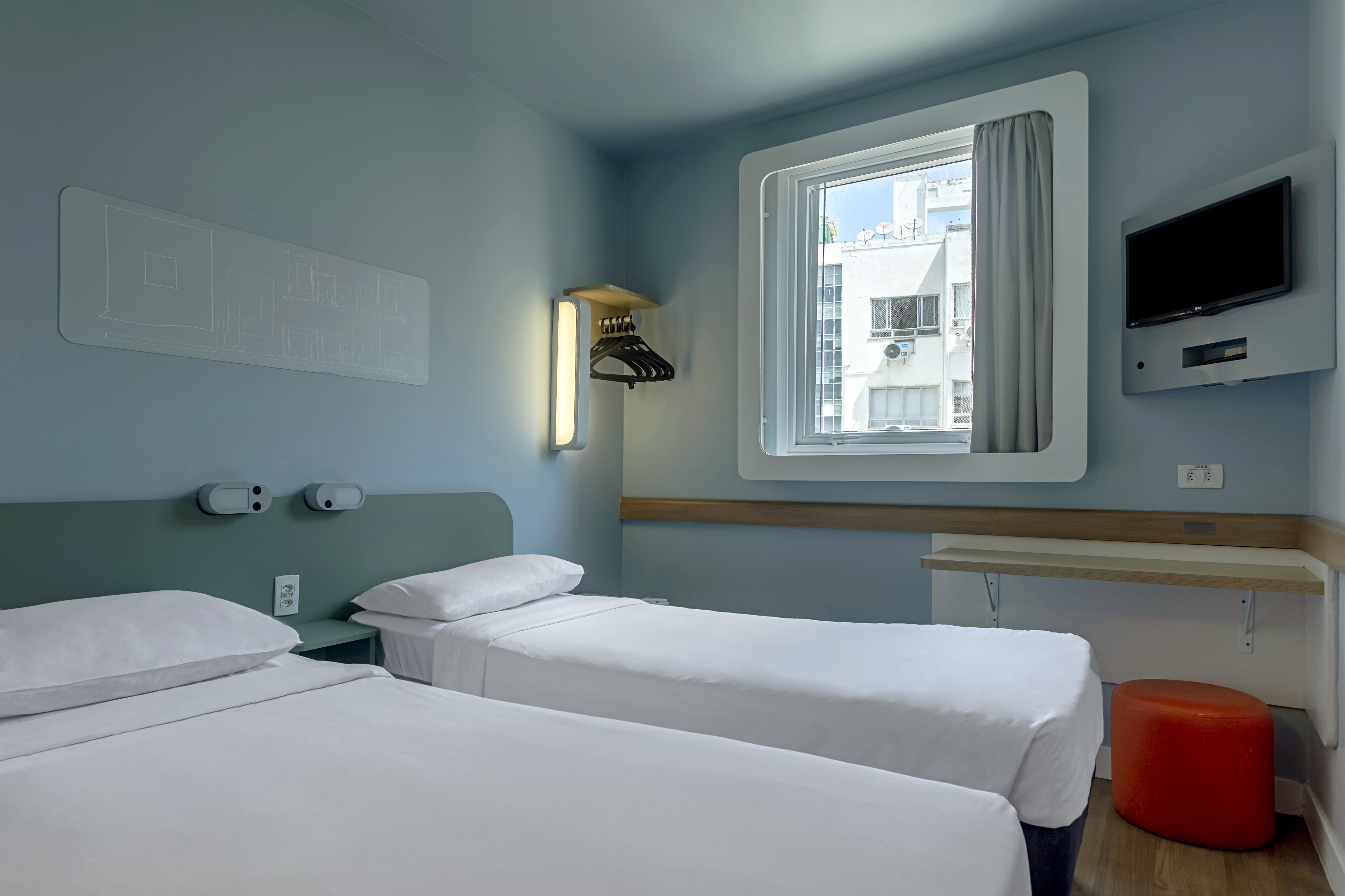 A hotel room with two beds, a TV, a window with a curtain, and a red ottoman.