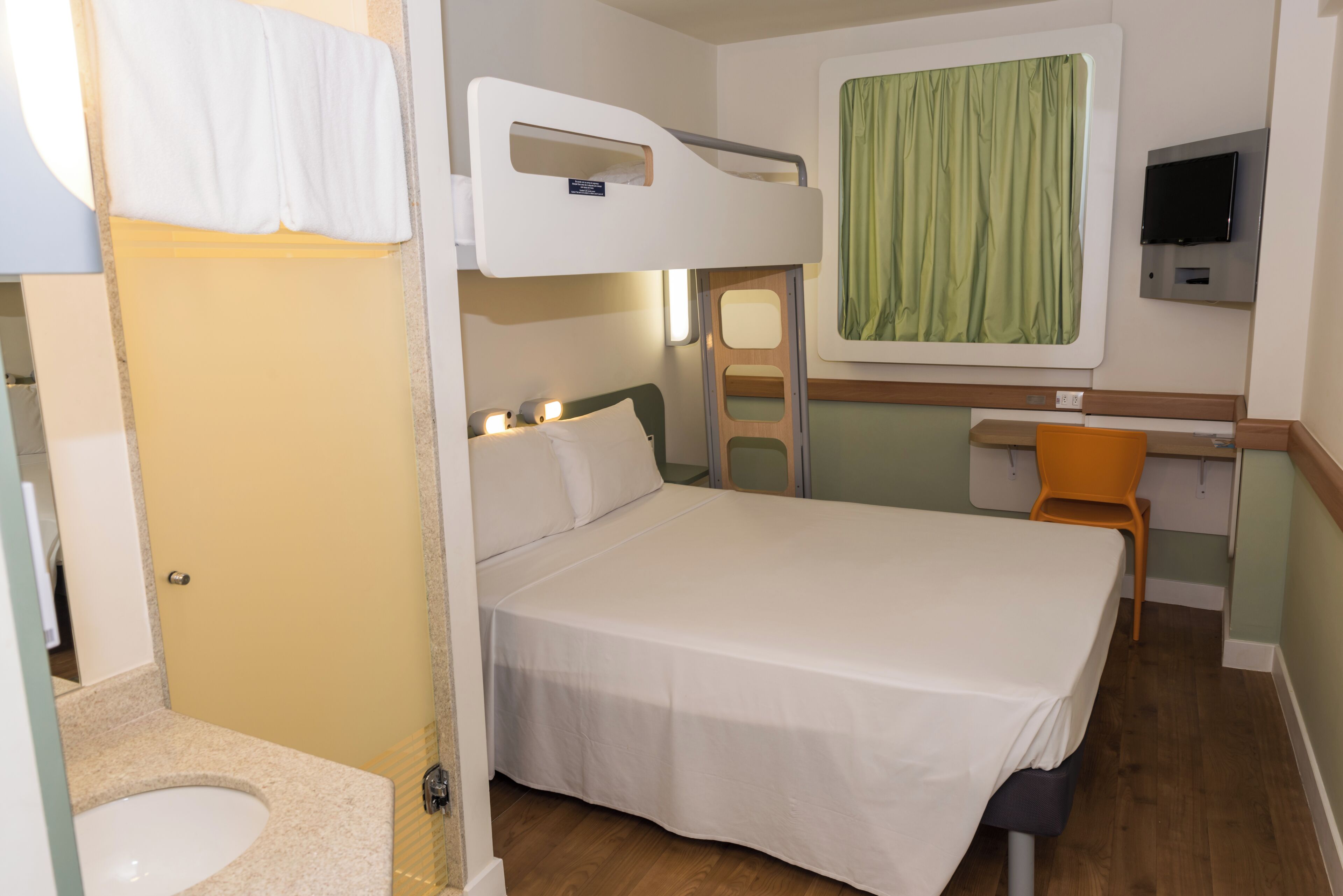 A compact hotel room with a bunk bed, a small desk with a chair, a television, and a window with curtains.