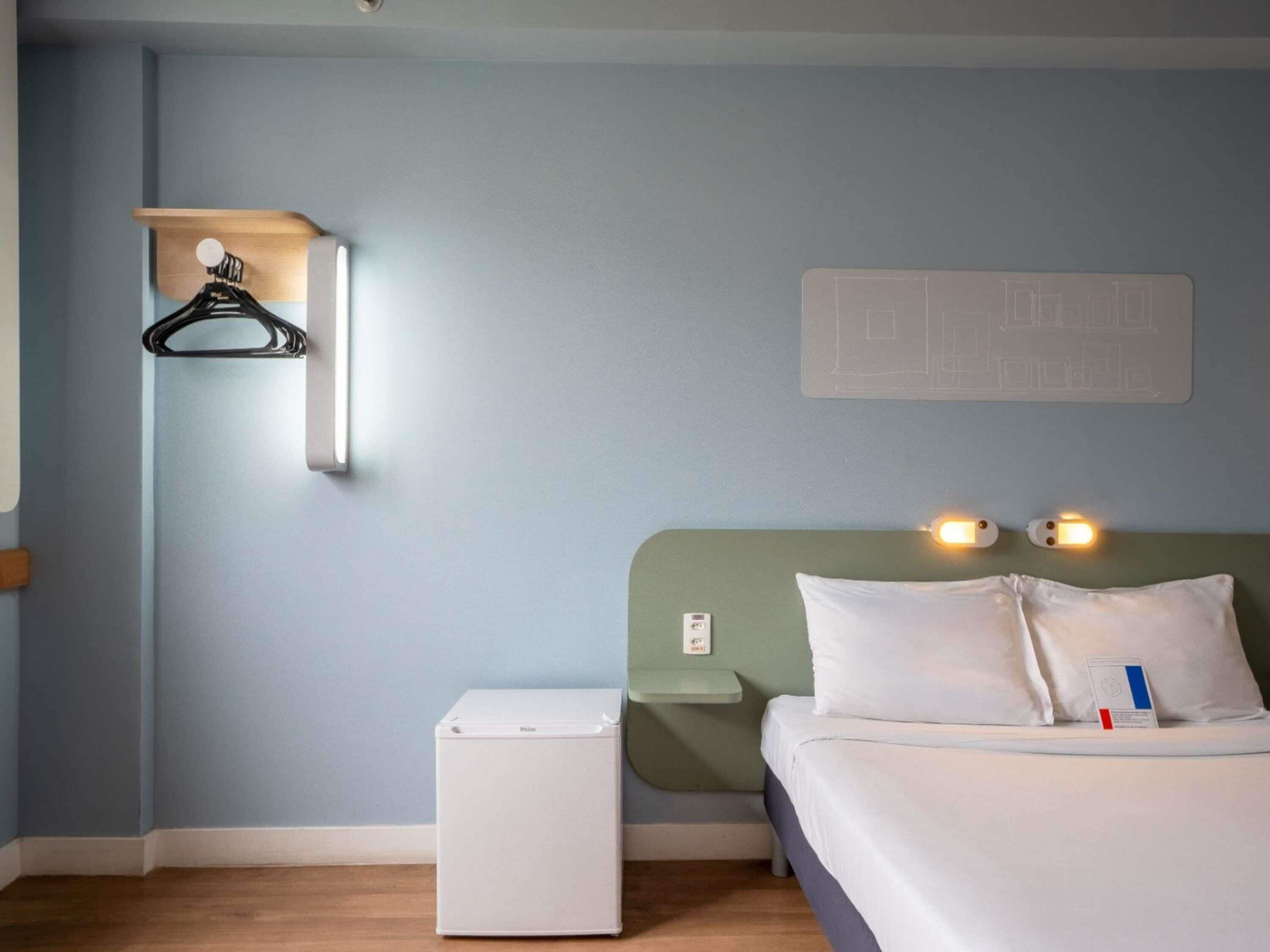 A hotel room with a bed, bedside table, a small white appliance, a wall-mounted light, and a wall decoration.