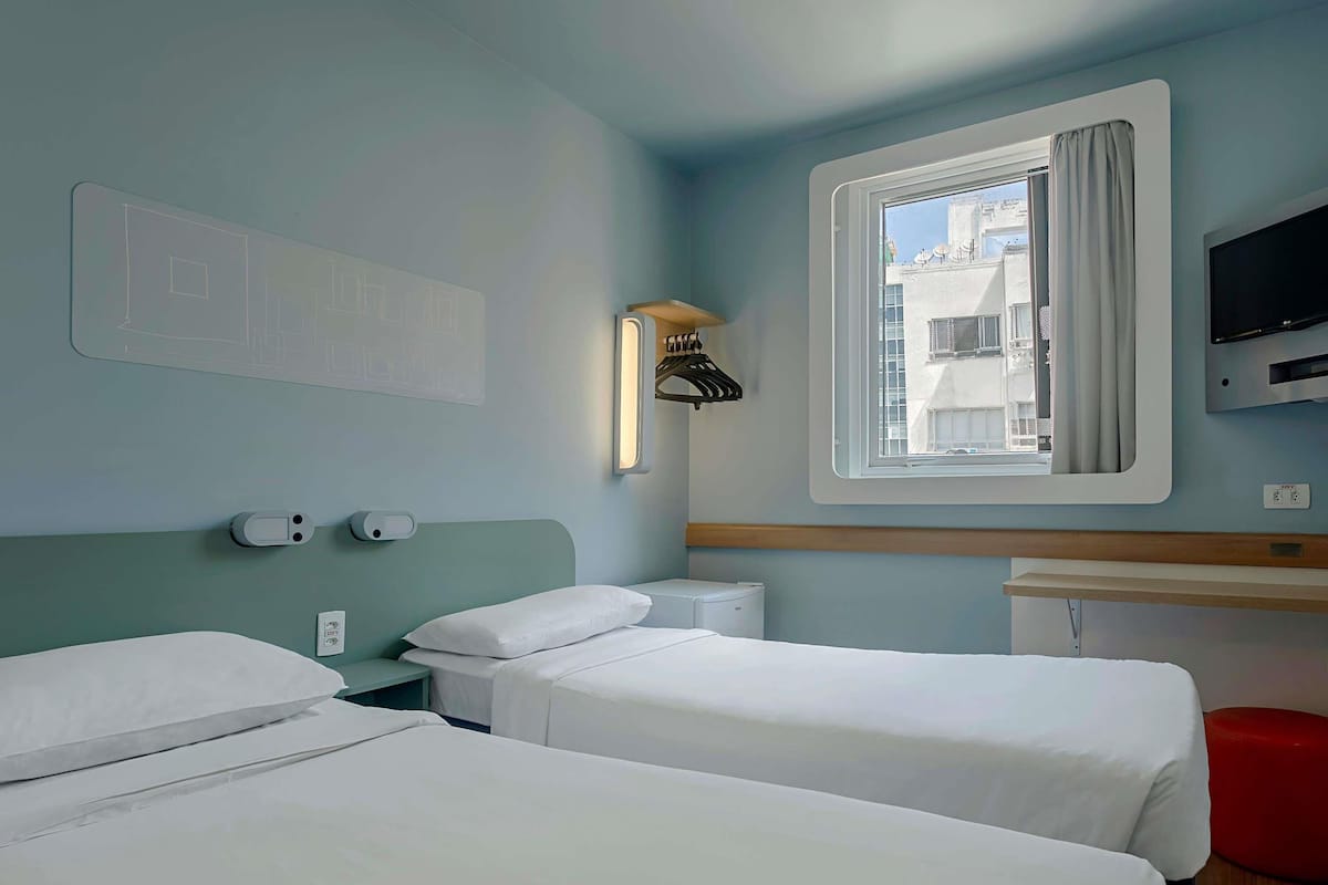 A hotel room with two beds, a TV, a window with curtains, a wall-mounted heater, and a wall-mounted light.