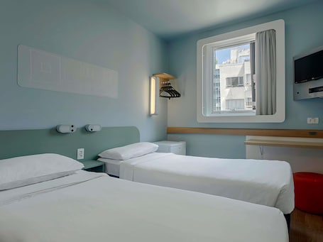 A hotel room with two beds, a TV, a window with curtains, a wall-mounted heater, and a wall-mounted light.. ibis budget RJ Praia de Botafogo