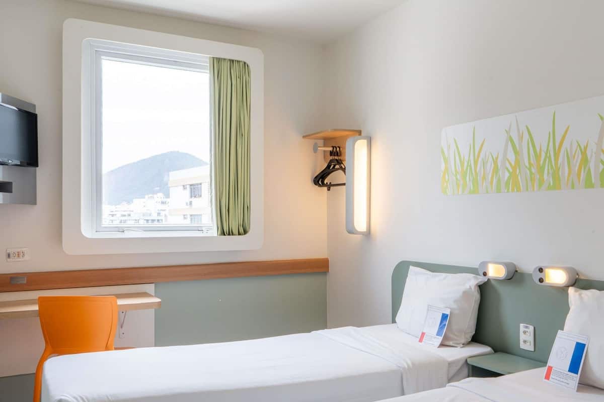 A hotel room with two beds, a desk, a TV, and a window with a view of buildings and hills.