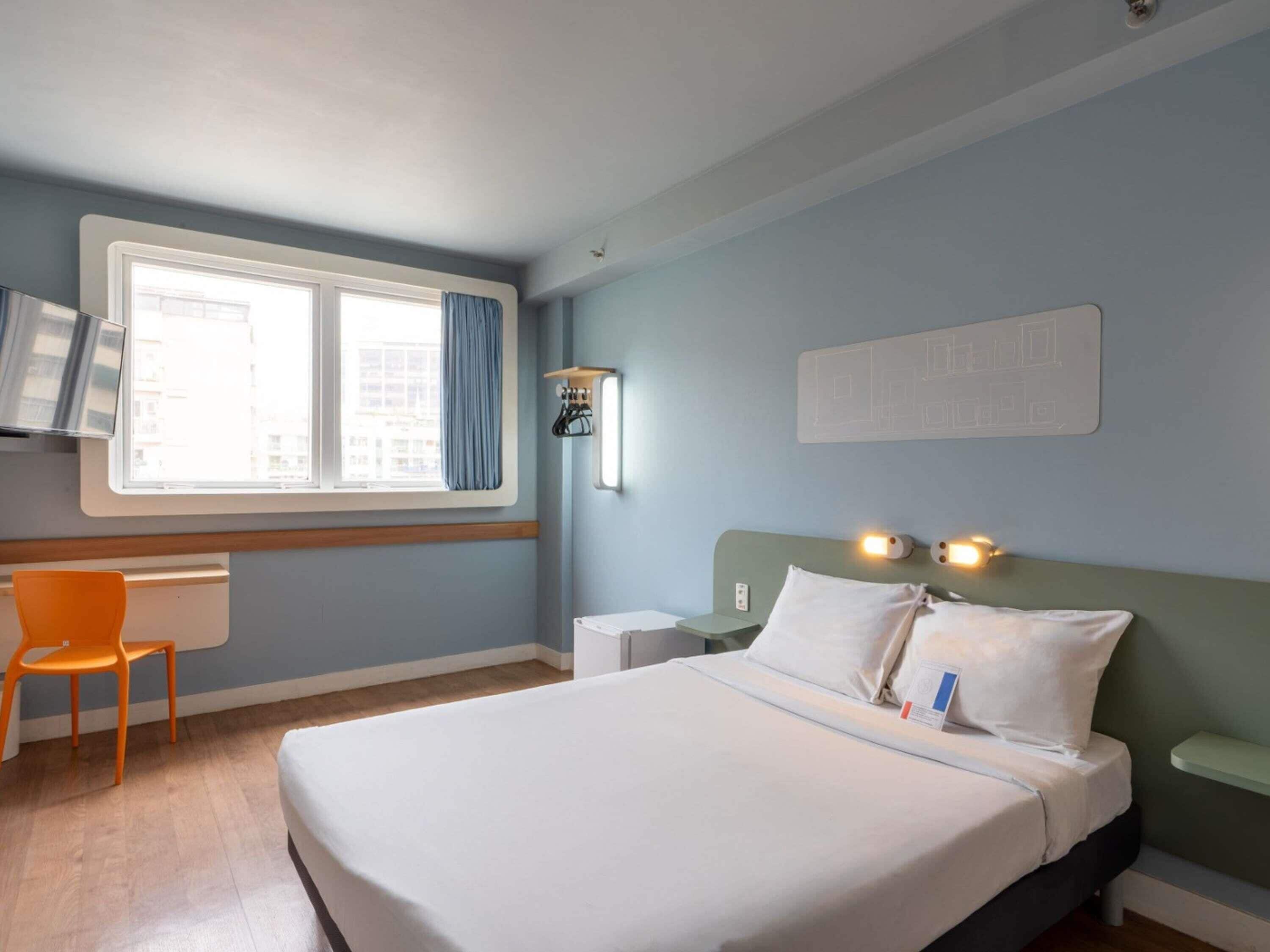 Una habitación de hotel con cama, escritorio, silla, ventana con cortinas y lámpara de pared.