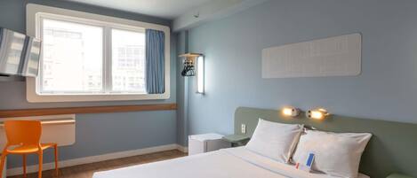 A hotel room with a bed, a desk, a chair, a window with curtains, and a wall-mounted light fixture.
