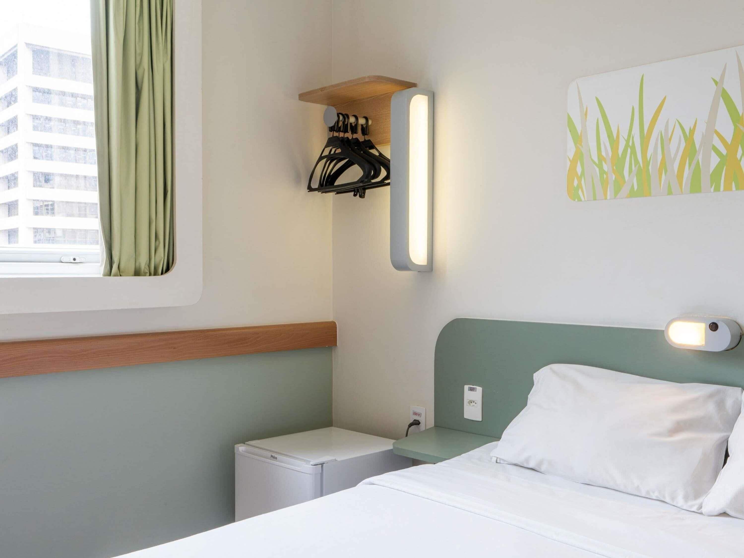 Hotelkamer met een bed, een klein nachtkastje, een wandlamp, een stopcontact en een raam met gordijnen.